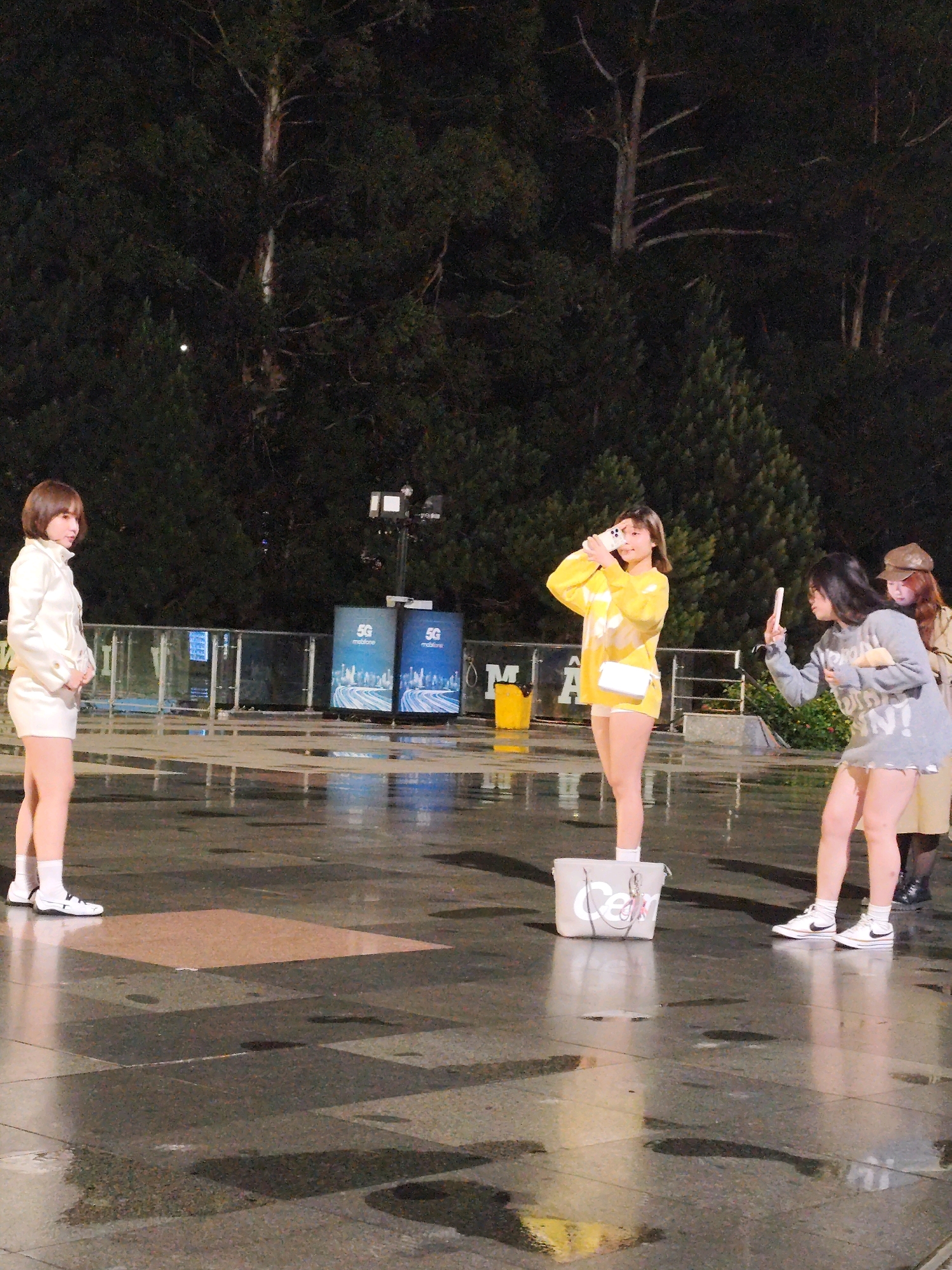 In the city of Da Lat, Vietnam, three young women are captured in an outdoor scene at night. The ground is wet and reflective, suggesting recent rain, and is paved with large square tiles. In the background, trees form a dark silhouette against the night sky, and two large screens displaying "5G mobifone" with images of a cityscape are visible.
The woman on the left stands facing away from the camera, wearing a white jacket and white shorts, with white socks and black-and-white shoes. She has short brown hair.
The woman in the center is facing the camera, holding a white smartphone up to take a picture. She is wearing a bright yellow long-sleeved sweatshirt with a white tie-dye pattern and white shorts, also with white socks and white shoes. A white purse hangs from her shoulder. She is standing on a light gray tote bag.
The woman on the right is facing the center woman, holding a white smartphone up to take a picture. She is wearing a gray sweater with white lettering and a light-colored skirt, with white socks and white shoes. Her hair is dark brown. Behind her, a woman in a brown hat and beige jacket is partially visible.
The overall scene appears to be a casual gathering, possibly at an outdoor event or public space, with an emphasis on smartphone photography.