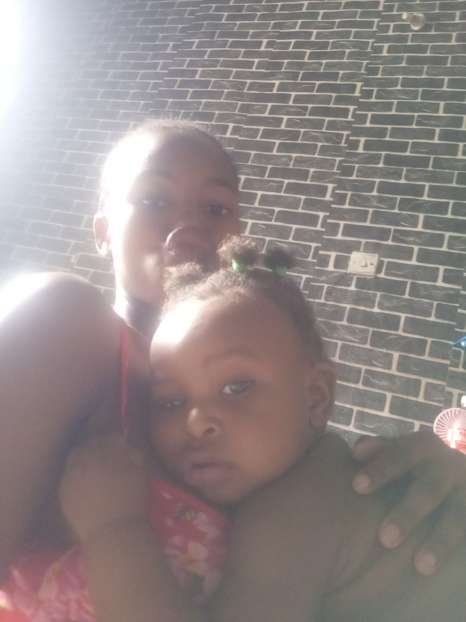 Here is a description of the image:
An adult and an infant are closely positioned together. The adult is taller and behind the infant, possibly holding them. The infant has curly hair with a small, green hair tie. They are both dark-skinned. Behind them, a brick-patterned wall with a light switch is visible. A small fan is partially visible to the right. Bright light is coming from the upper left of the image, washing out details in that area. The specific location and city are not discernible.