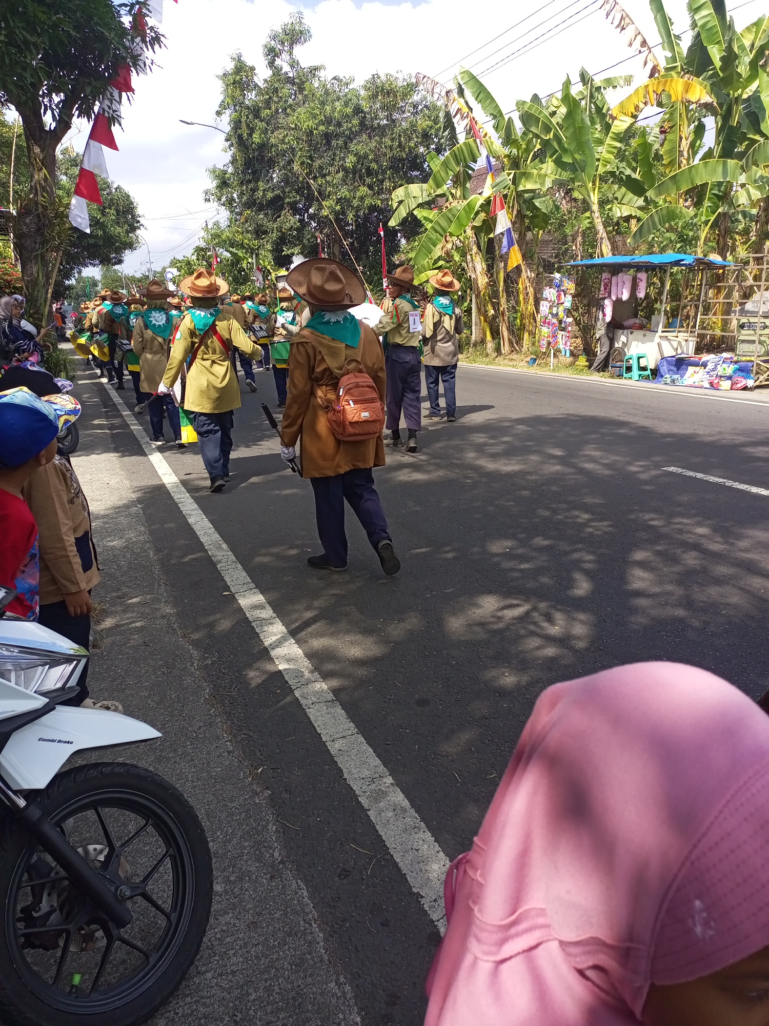 In Nanggulan, Indonesia, a group of young people in scout uniforms participate in a parade on a sunny day. They are walking on a paved road lined with trees and banana plants. Some individuals wear traditional wide-brimmed hats and carry backpacks. The parade features red and white flags, possibly indicating a national celebration. To the right of the road, a small roadside stall displays various items for sale. In the foreground, a white motorcycle is parked, and a person wearing a pink hijab is visible. The scene captures a vibrant community event within a tropical, rural setting.