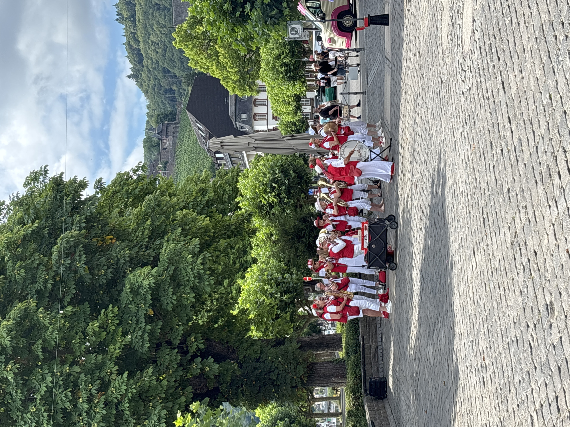 In Cochem, Germany, a brass band is playing in a cobblestone plaza. The band members are wearing red vests over white shirts and white pants. They are holding and playing various brass instruments, including saxophones, trumpets, and tubas. Some members are also holding drums. A red and white wagon is placed in front of some of the band members. In the background, there are trees, buildings, and a hill with a castle on it. A pink and white vintage van is parked in the background to the right.