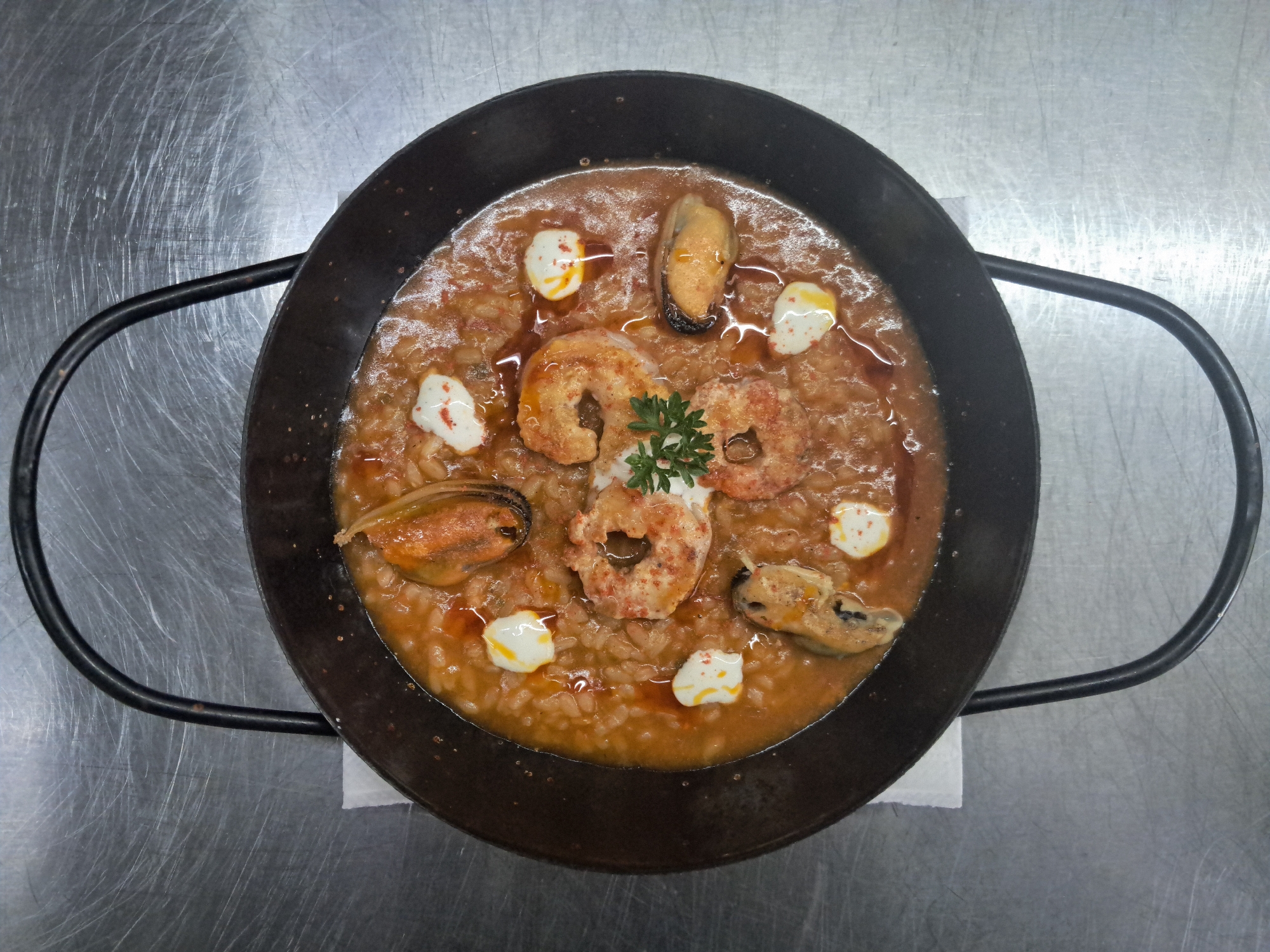 A close-up, high-angle shot showcases a paella dish in a black pan. The paella consists of rice, seafood, including shrimp and mussels, garnished with dollops of a white creamy substance sprinkled with red pepper flakes, and a sprig of parsley. The pan has two black handles, and rests on a white napkin. The backdrop is a textured, brushed stainless steel surface. The image was taken in Vicente López, Argentina.