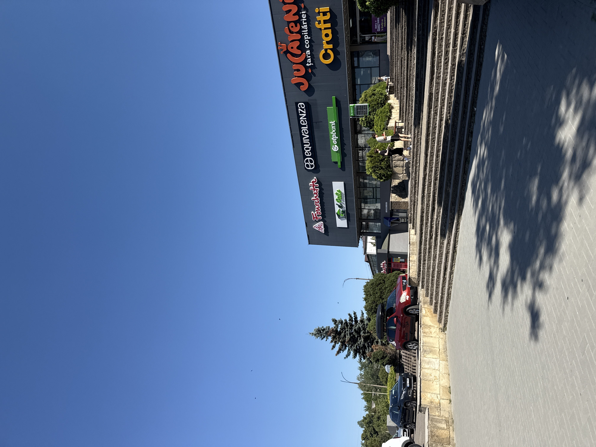 The image shows a modern shopping center facade under a clear blue sky. Several businesses are visible on the building's exterior, including "Fourchette," "Ekvivalenza," "otpbank," "Jucărenii - țara copilăriei," and "Crafti." The shopping center is elevated, with a wide set of stone stairs leading up to its entrance. In front of the building, there are various green bushes and trees. In the foreground, a paved area with brick-like patterns is visible, with shadows cast by nearby trees. To the left of the shopping center, there are parked cars, including a dark-colored SUV and a red SUV with a roof carrier. A tall evergreen tree stands between the parked cars and the shopping center. The location is identified as Bălți, Moldova. Two young men are walking up the stairs, and a woman is visible further up the stairs near the entrance. A banner with Romanian text is displayed at the entrance.