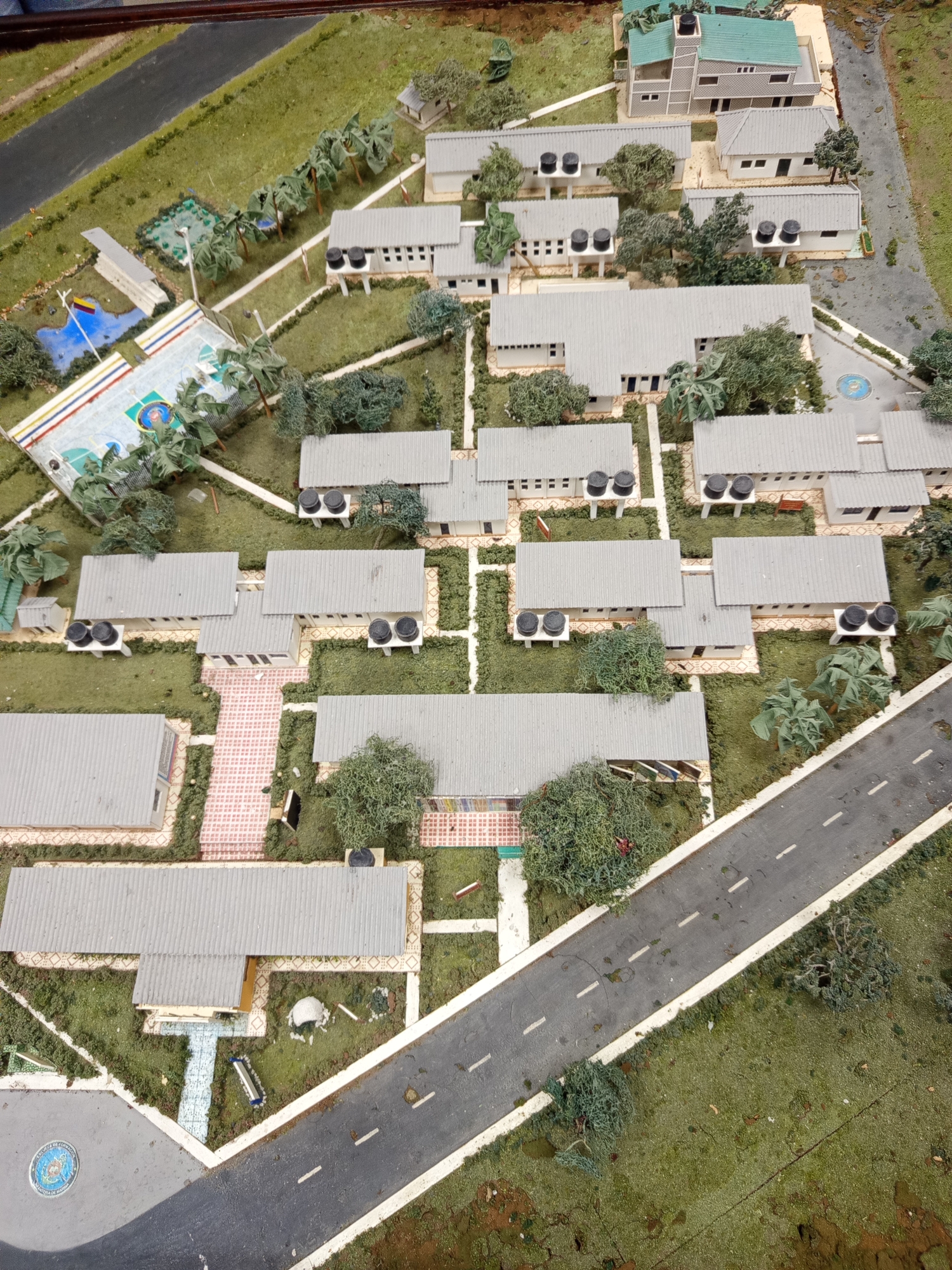 This is a high-angle, medium shot of a model of a facility in the city of Coveñas, Colombia. The model depicts a complex of single-story, rectangular buildings with gray, corrugated metal roofs. The buildings are arranged around courtyards and are connected by paved walkways and roads. Each building appears to have multiple windows and doors, and some have air conditioning units mounted on the roofs.
The surrounding environment is meticulously detailed, with miniature trees, grass, and a road. In one section of the model, there is a sports court with painted lines and palm trees. There is also a small blue pond with a flag representing Colombia. The road in the model has white dashed lines and appears to be bordered by grass and some scattered debris. At the entrance to the facility, there is a circular emblem that reads "POLICIA DE COMANDO". The model is placed on a textured surface that resembles ground, with some patches of darker brown, possibly representing soil or dirt.