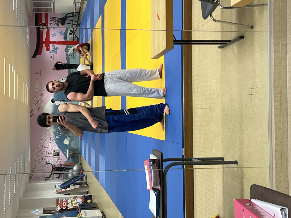 Two men stand on a blue and yellow judo mat, reflected in a mirror. One wears a black shirt and white pants, the other a gray shirt and blue pants. The second man holds a phone, seemingly taking a selfie. In the background, a wall features a mural with Japanese-inspired art, while workout equipment is visible along the wall. A table and some office supplies are also in the shot.