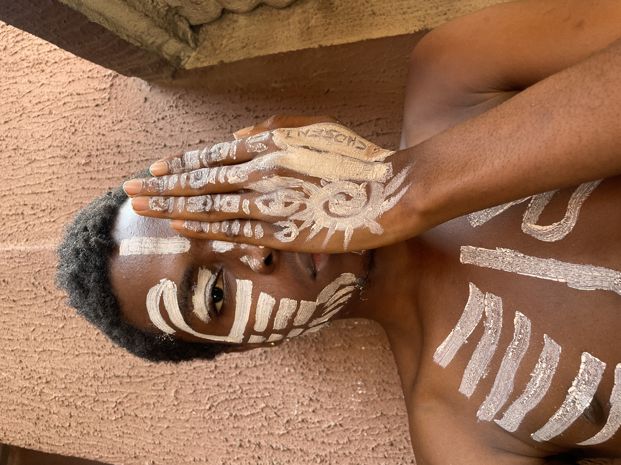 A person, appearing to be male, with dark skin and short, curly black hair, is captured in a close-up, artistic shot. They are lying on their side, bare-chested, against a textured, reddish-brown wall or ground surface, likely in Onitsha, Nigeria. The person's face, hand, and torso are adorned with striking patterns of white and light beige body paint, evoking tribal or ceremonial markings.
The individual's left hand is raised, partially covering their face and forehead. On the back of this hand, a prominent white design resembling a stylized sun or spiral is painted. On the wrist area, the word "CHOSEN" is visible in black capital letters, written vertically on a light beige paint patch. Bold white paint strokes create geometric patterns on the face, particularly around the visible right eye and cheek. The chest features similar white striped patterns, hinting at a ribcage-like design.
Bright, natural lighting illuminates the scene, casting a shadow from an unseen structure, suggesting the photo was taken during the day. The person's visible eye is open and looks directly at the viewer with a calm, focused expression, conveying a sense of introspection or a deliberate artistic presentation.