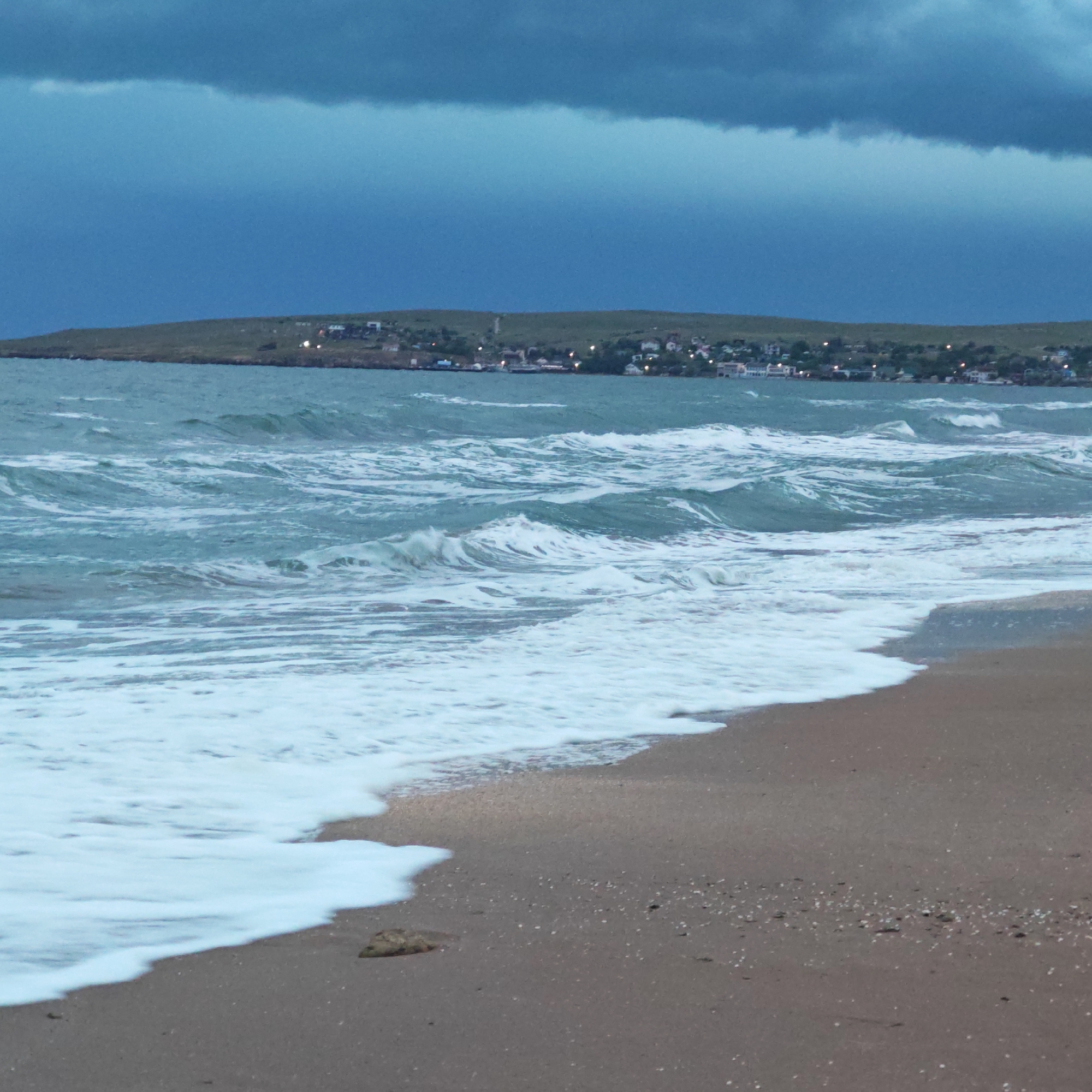 The image shows a beach scene in городское поселение Щелкино, Ukraine. The foreground features a sandy beach with visible shell fragments. White foamy waves are breaking on the shore. In the background, the sea is choppy with visible waves. Further back, a low-lying landmass with buildings (likely a town or settlement) is visible. The sky is overcast with dark, heavy clouds. The lighting suggests either early morning or late evening. There are no people visible in the image.