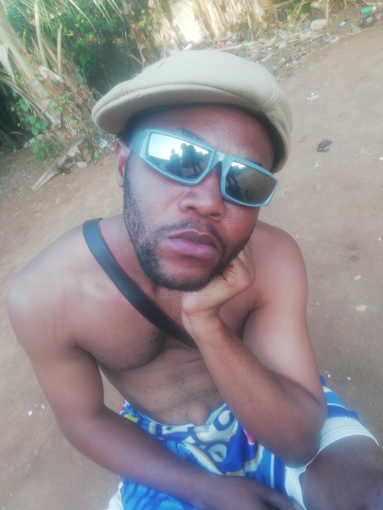 A low-angle, medium shot captures a bare-chested man with brown skin. He sports a dark beard and is wearing sunglasses and a beige cap. He has a black strap across his shoulder and is draped in a blue and white patterned fabric. His left hand is positioned against his cheek, suggesting a contemplative pose. The environment appears to be an outdoor setting, potentially a yard or open area, with dirt or sand visible on the ground. Some greenery and debris can be seen in the background. The location is the city of Unknown, Unknown.