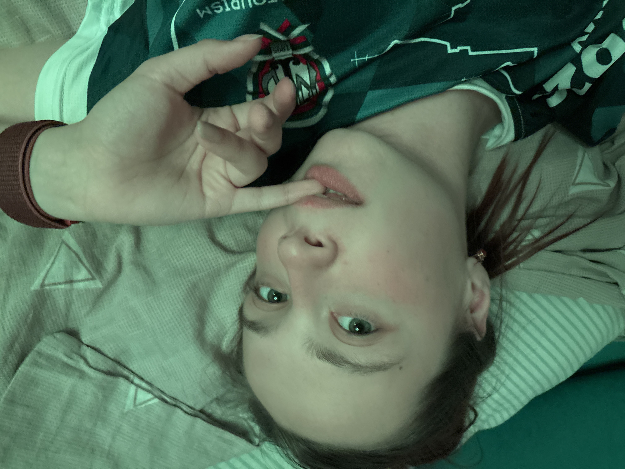 A young woman lies on a bed covered in a striped blanket and a patterned pillow, wearing a dark green jersey with a white collar. She has light skin, green eyes, and her hair is pulled back, with some strands loose around her face and right ear. Her left hand is raised with the index finger touching her lips. A red-brown wristband is visible on her left wrist. The location cannot be determined from the image.