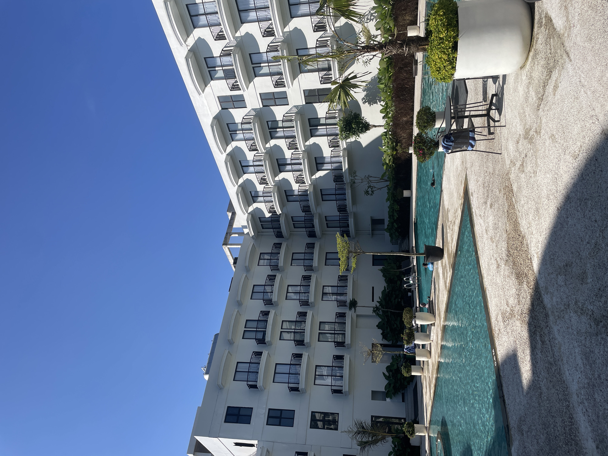 This image depicts an outdoor swimming pool area in Kayuambon, Indonesia, situated in front of a multi-story white building, likely a hotel or resort, under a clear blue sky. The building features numerous balconies with black wrought-iron railings and distinctive curved white canopies above dark-framed windows, repeated across at least four visible floors. Strong sunlight casts sharp shadows from the building and vegetation onto the light-colored, textured paving around the pools.
Two interconnected or adjacent swimming pools are present, filled with clear turquoise water. One section of the pool has a depth marker of "0.6 M". Inside the pools, at least two individuals are visible: one person partially submerged and another on a blue inflatable ring.
The poolside area includes various elements: large white planters containing green foliage, smaller trees directly planted in the ground, and a dense, dark green planted bed along the base of the building. A small dark table with two dark chairs is positioned on the right foreground, with a blue and white striped towel draped over one chair. A palm tree is also visible near the foreground. A pool ladder is visible in the further pool section.