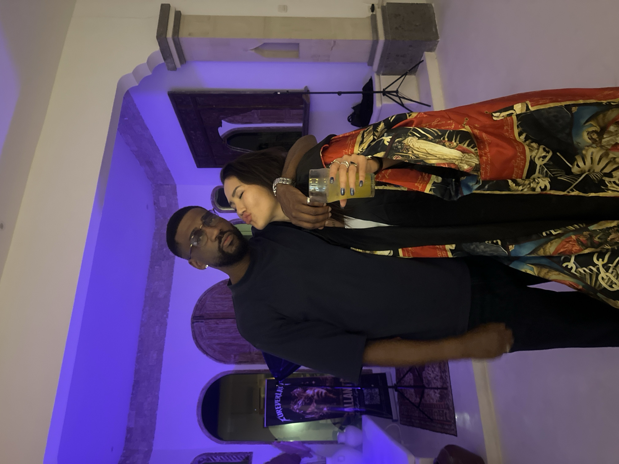 A Black man wearing glasses and a dark t-shirt embraces a woman who is wearing a patterned robe. The woman has her lips pursed as if to kiss the man, and she is holding a drink with a straw. The man has his arm around her. They appear to be indoors in a room with purple lighting. There is a poster in the background and a tripod with a light. This scene is located in the city of North Kuta, Indonesia.