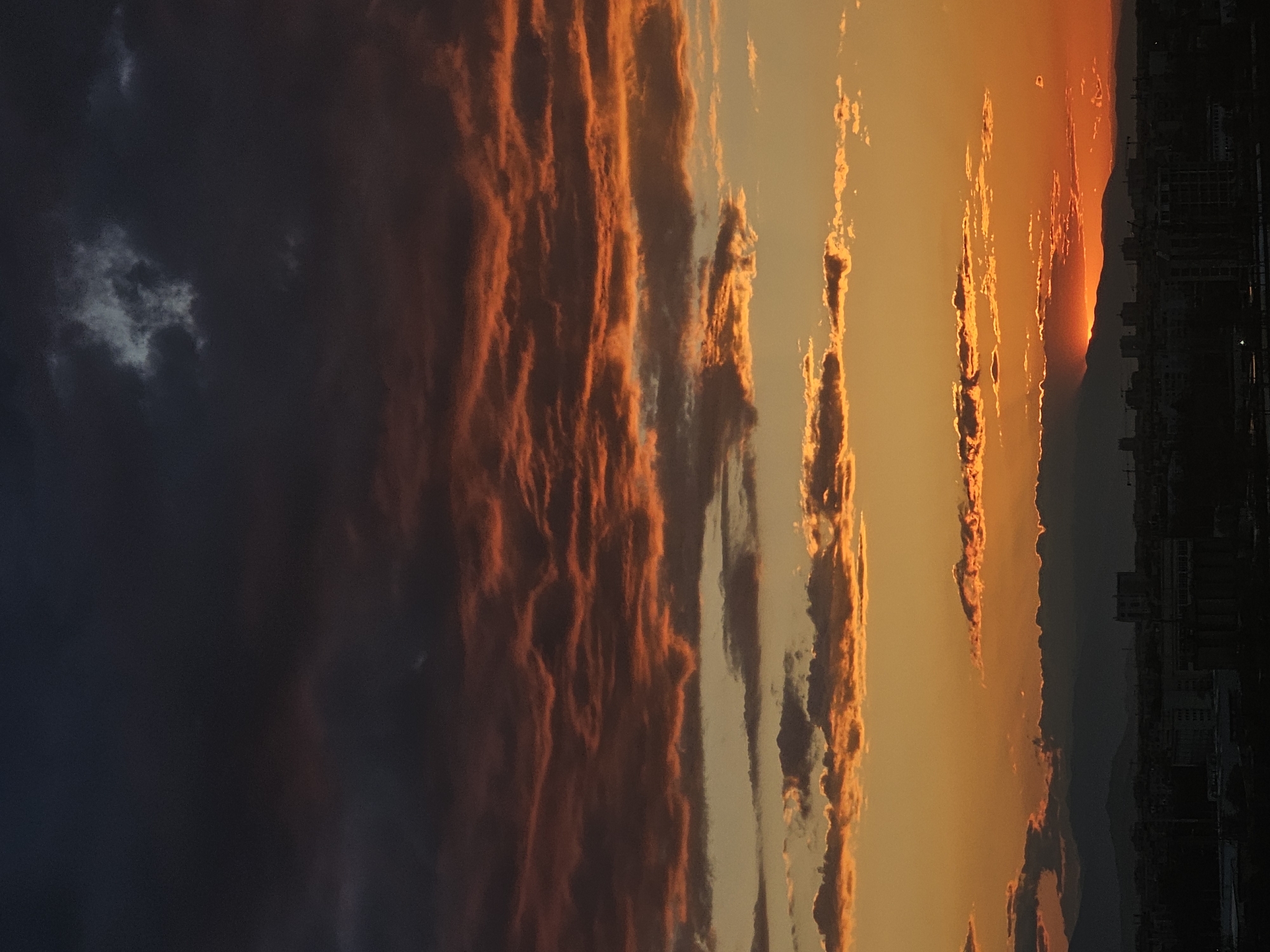 The image depicts a sunset over Plovdiv, Bulgaria. Striated orange and red clouds dominate the sky, transitioning from lighter hues near the horizon to darker shades on the left. The silhouette of a distant mountain range is visible against the bright sky, with the sun appearing to set behind it. The skyline of Plovdiv is faintly visible in the foreground, composed of buildings and structures. The photograph is taken from an elevated position, providing a wide view of the city and its surrounding landscape.