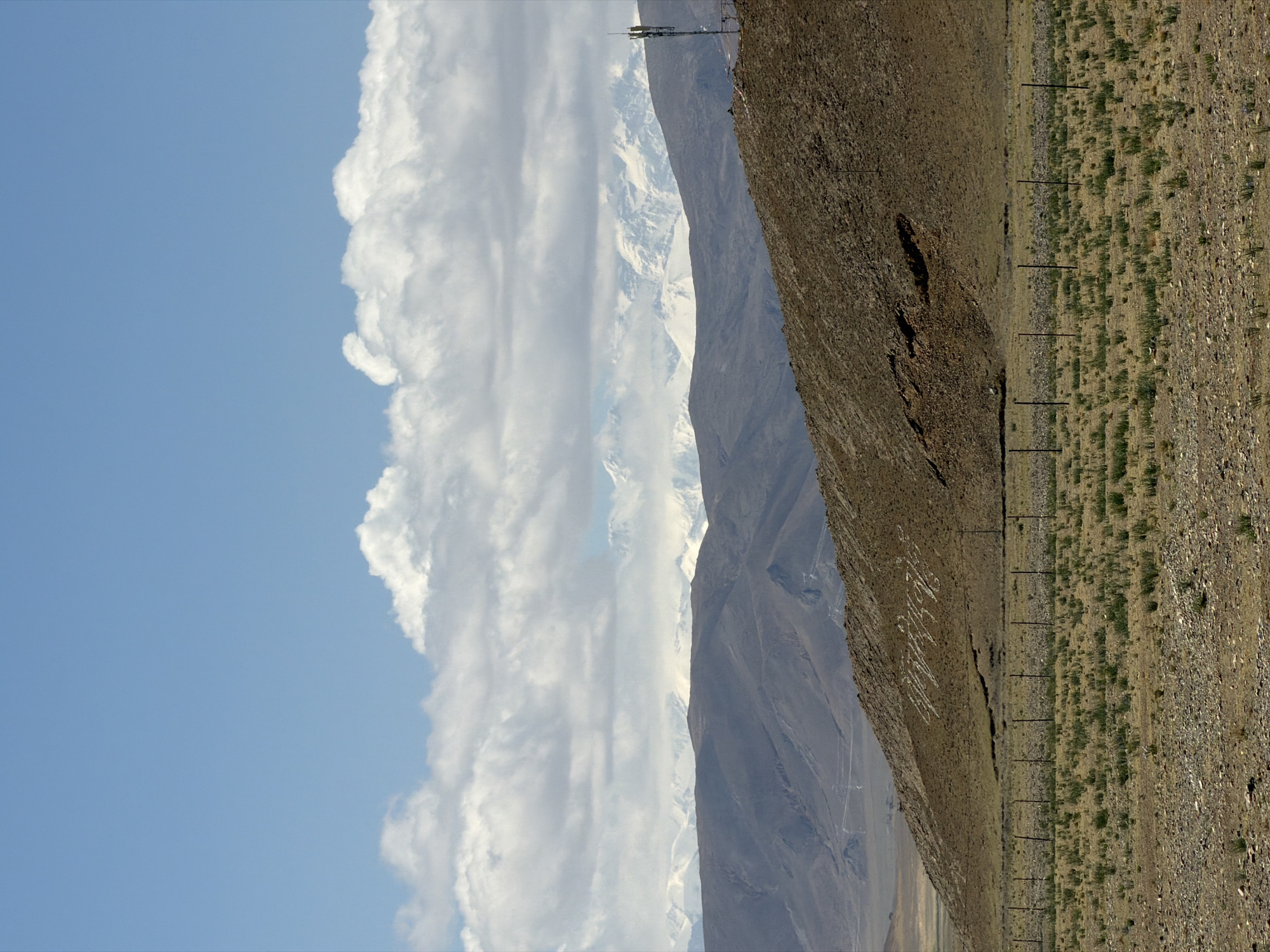 This image captures a vast, arid mountainous landscape under a bright, partly cloudy sky. The scene depicts a remote, high-altitude environment, likely in Mainpu township, China, as suggested by the location context.
In the upper portion of the frame, a clear blue sky is visible, adorned with scattered, fluffy white cumulus clouds. Below the sky, in the mid-ground, a prominent range of majestic mountains rises, their peaks covered in snow, creating a striking contrast with the darker, rocky slopes. The distant mountains appear somewhat hazy.
The foreground is divided into two main areas. To the right, there is a steep, rugged hillside composed of brown and grey rock and earth. On this hillside, a large inscription in white characters is clearly visible. The text reads "我爱中华" (Wǒ ài Zhōnghuá), which translates to "I love China." To the left of this hillside, a relatively flatter, rocky plain stretches out, sparsely covered with tufts of dry, light green and yellow vegetation. A series of dark, thin fence posts runs horizontally across this plain, suggesting a boundary or infrastructure line.
No direct human activity is taking place. The overall impression is one of a grand, rugged natural landscape, marked by human presence through the fence and the large patriotic inscription. A tall, slender structure, possibly a communication tower or weather mast, can be seen on a distant peak. The lighting suggests bright daylight, likely late morning or early afternoon, with clear visibility.
