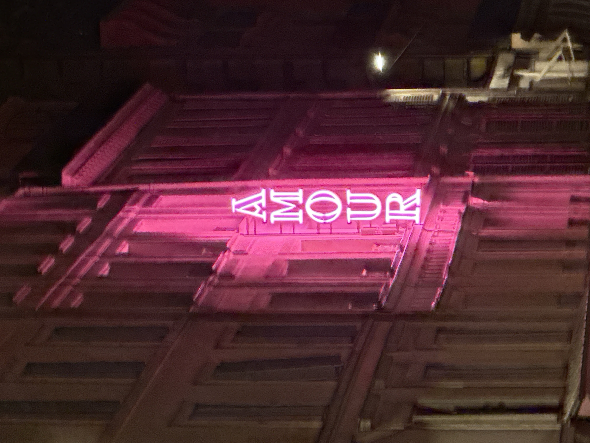 The image shows a building facade with a neon sign that reads "AMOUR" in pink letters. The architectural style appears to be art deco. The location is likely Paris, France, considering "Amour" is French for "love" and the building's design is consistent with Parisian architecture. The image seems to be taken at night, with a single light source visible in the upper part of the frame.