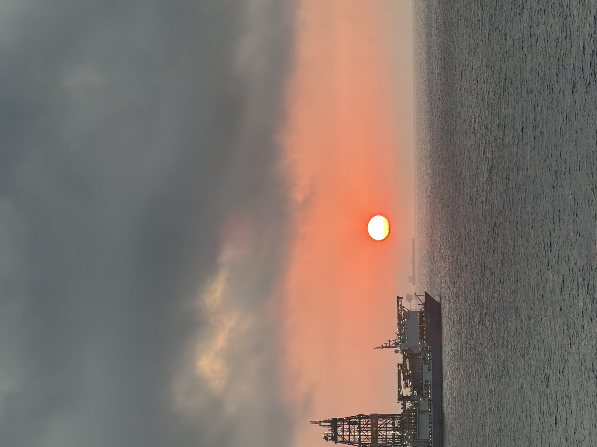 The image captures an expansive seascape at either sunrise or sunset, appearing to be rotated 90 degrees counter-clockwise from its natural orientation. If viewed correctly, the horizon runs horizontally across the middle of the frame.
The sky, occupying the upper portion of the scene, displays a dramatic contrast. On the left, heavy, dark grey clouds dominate, gradually transitioning towards the right into a luminous expanse of vibrant orange and reddish hues. A brilliant, circular sun, glowing intensely white at its core and orange around its edges, is prominent on the right side of the sky, near the horizon line.
Below the horizon lies a vast body of water, likely an ocean or sea. The water's surface is dark grey, textured with visible ripples and waves, subtly reflecting the warm light from the sky.
In the foreground, positioned in the lower right section of the image (when oriented correctly), is a large, complex industrial vessel or offshore platform. Its intricate structure, featuring multiple decks, cranes, and scaffolding, is silhouetted against the glowing sky. Several artificial lights are visible on the vessel, suggesting ongoing operations or the transition into twilight. Faintly in the distance, further across the water, the silhouette of another, smaller vessel or structure can be observed.
The time of day is clearly either sunrise or sunset, indicated by the low position of the sun and the rich, warm colors in the sky, creating a serene yet industrious atmosphere. There are no people or discernible text visible in the image.
