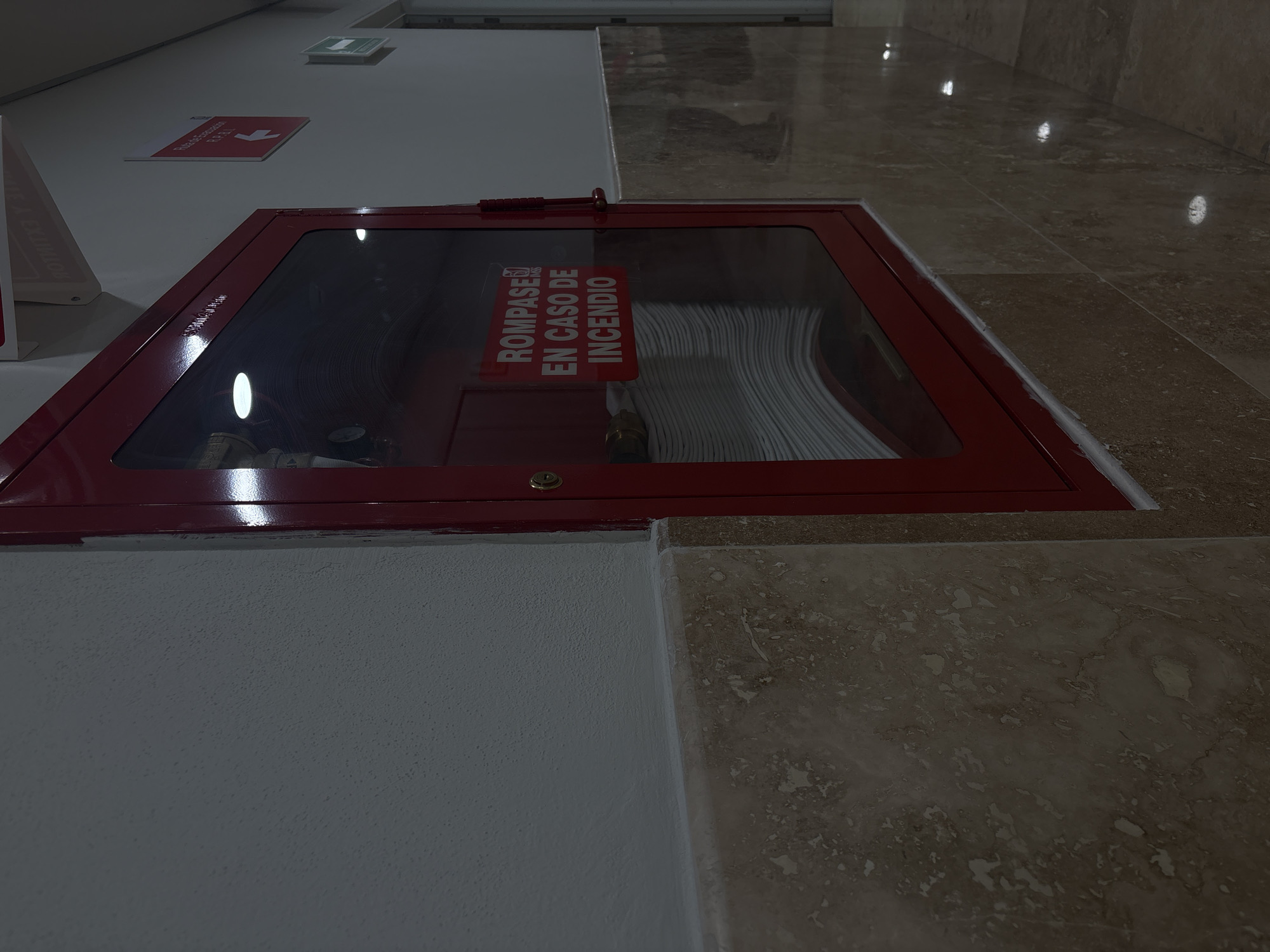 The image shows a fire hose cabinet recessed into a wall with a marble baseboard. The cabinet is red and has a glass front with a sign in Spanish that reads "ROMPASE EN CASO DE INCENDIO" (Break in case of fire). Inside the cabinet, a coiled fire hose is visible. The wall is white, and the floor is tiled. The location is likely a public building in Puebla, Mexico, given the Spanish signage and architectural style. There are no people visible.