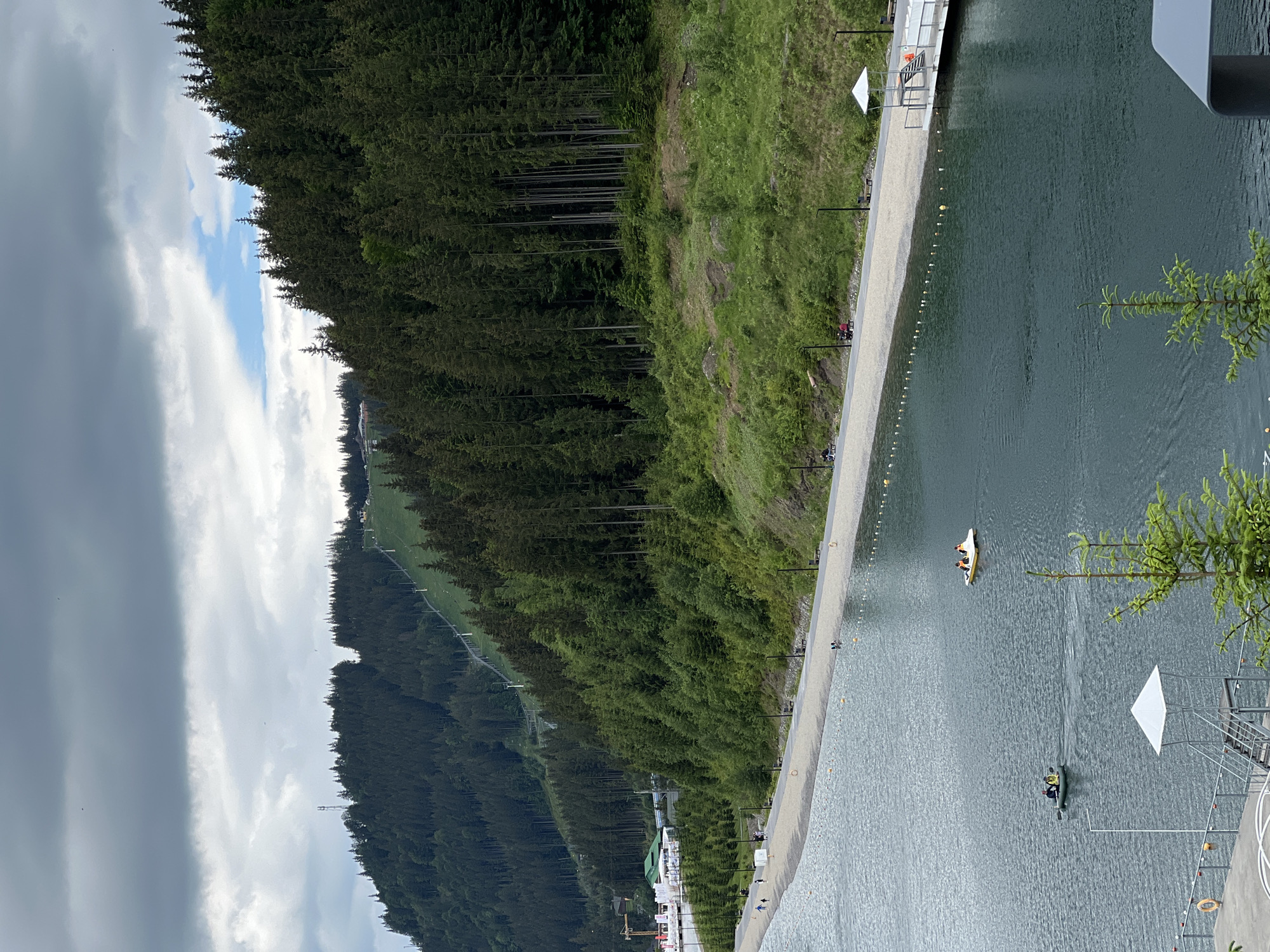 An elevated, medium shot captures a wide, dark body of water with a sandy shore and a treeline beyond. On the water, two small boats, one a pedal boat with two people, and another a small inflatable boat with two people, are visible. Along the sandy shore, a paved path is lined with lampposts and benches. People can be seen walking and sitting along the path, and a lifeguard tower with a white umbrella stands on a wooden pier. The background is dominated by a steep, tree-covered hillside with a ski slope visible near the top. The sky is overcast with dramatic clouds.