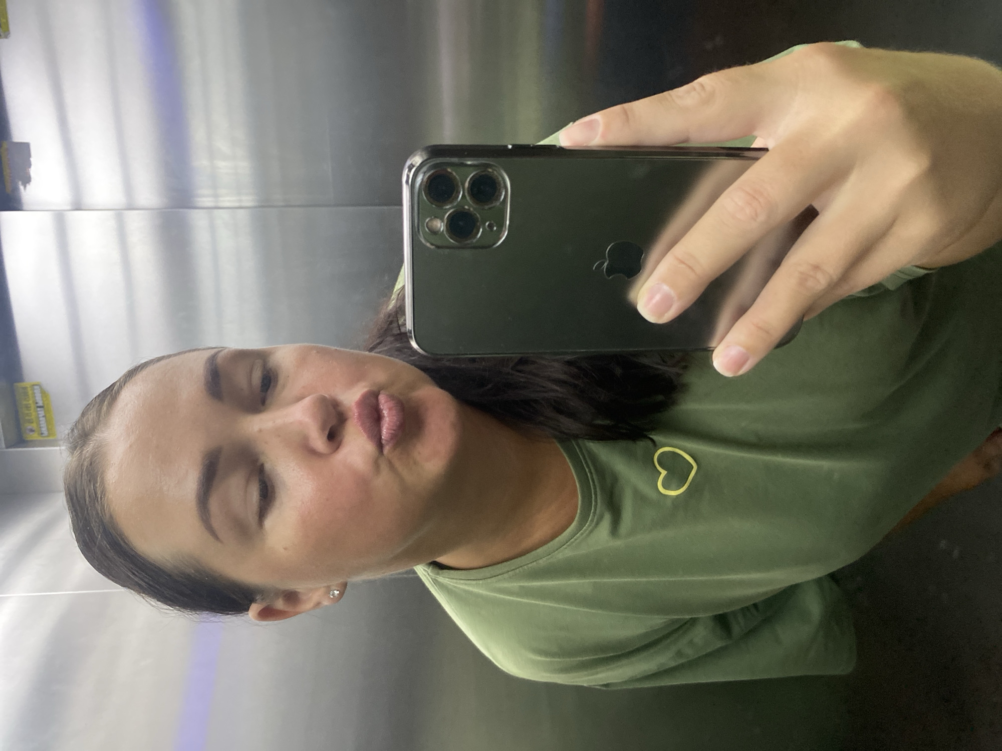 A young woman with dark hair pulled back takes a selfie in an elevator. She is wearing a green t-shirt with a yellow heart design on the left chest. She is puckering her lips. She is holding a dark-colored smartphone in her right hand, which is reflecting the elevator's metallic walls. The elevator appears to be in a modern building. The context provided suggests the photo was taken in Sochi, Russia.