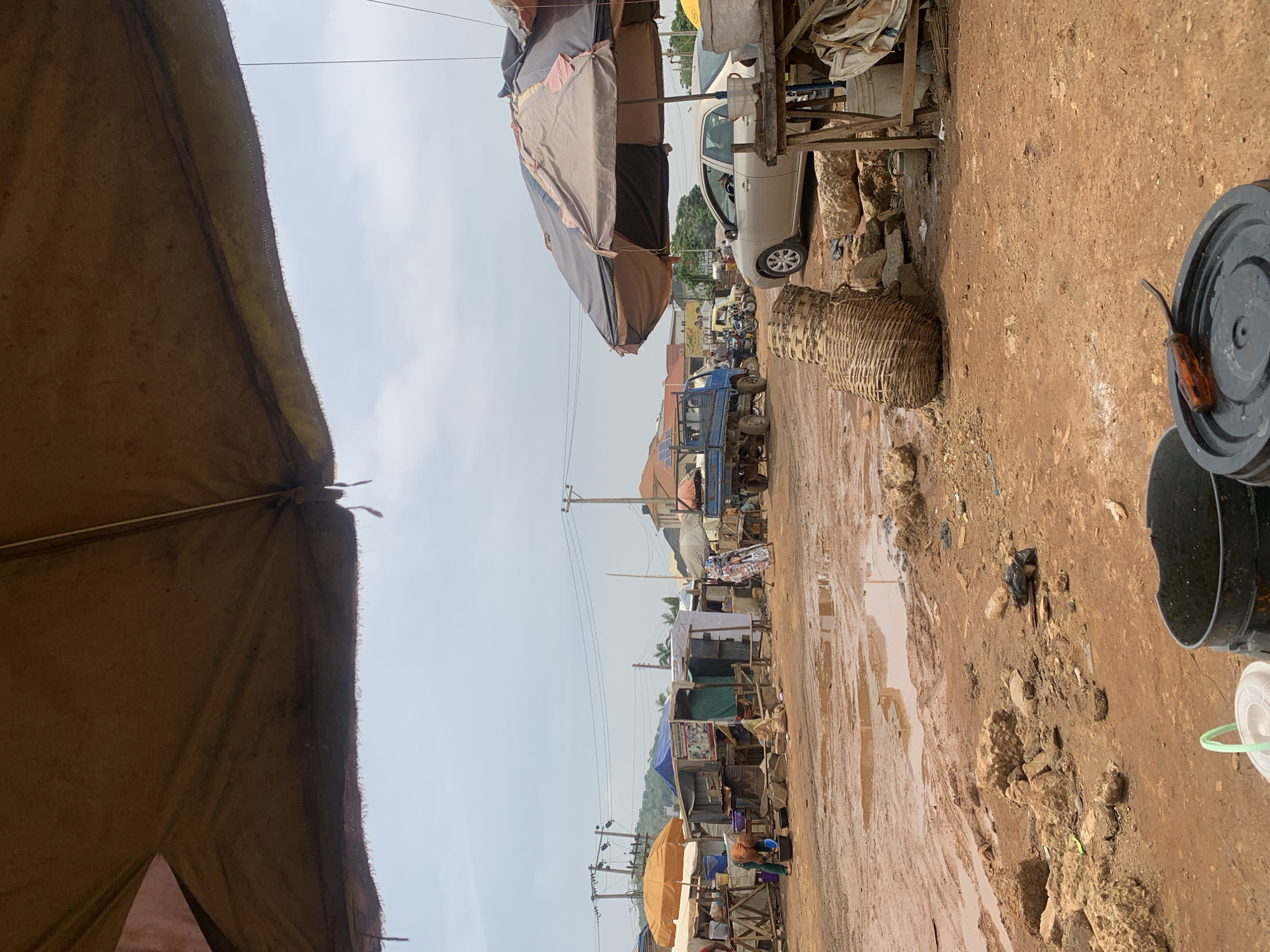 The image depicts a muddy street scene in Dutsen Alhaji, Nigeria. The road is unpaved, saturated with water, and littered with debris. Temporary stalls and vendors' structures line both sides of the street, covered by tarpaulins and umbrellas. Vehicles including cars and trucks, along with motorcycles are visible further down the road. Baskets and buckets are positioned on the side of the road in the foreground. Power lines and utility poles run along the street, indicating basic infrastructure. People are sparsely visible among the stalls. The overall impression is one of a busy, somewhat disorganized marketplace environment.