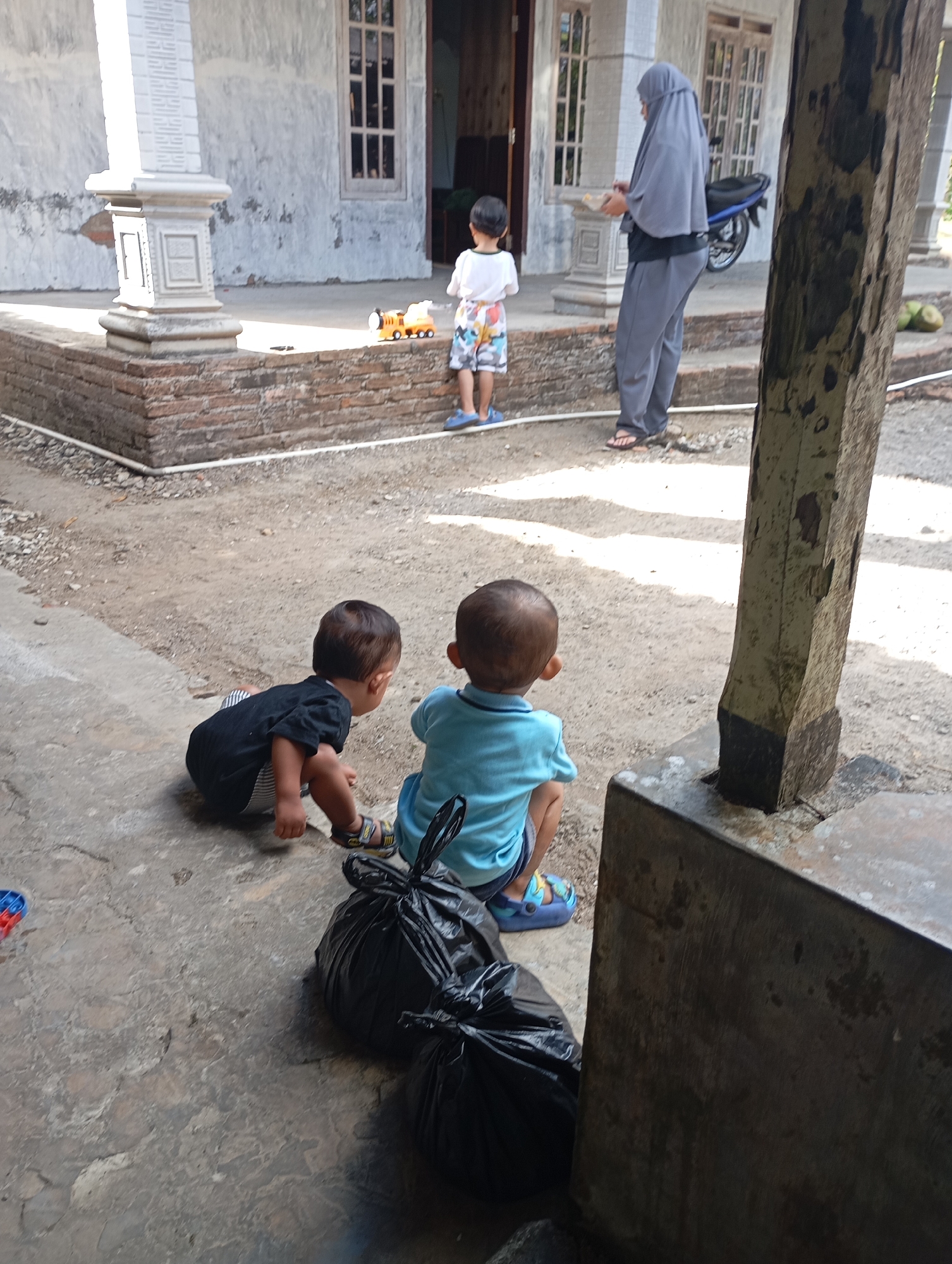 In Ketepung, Indonesia, two young children are observed from behind, seated on a concrete surface in what appears to be a residential yard. The child on the left, wearing a black t-shirt and striped shorts, is in a crouched position, facing towards the right. The child on the right wears a light blue t-shirt and patterned shorts, also facing away from the viewer. Both children are wearing sandals.
In the background, a woman in a grey hijab and matching pants stands near the entrance of a building, holding a plate. She is facing the children. Another young boy in a white t-shirt and patterned shorts stands a short distance away, near a toy train. The building behind them has a white facade with large windows and pillars. A motorcycle is parked to the right of the building entrance. The ground is a mix of gravel and concrete, with some vegetation visible. Two black garbage bags are positioned in the foreground, near the seated children. The scene is lit by natural daylight, suggesting it is outdoors during the day.