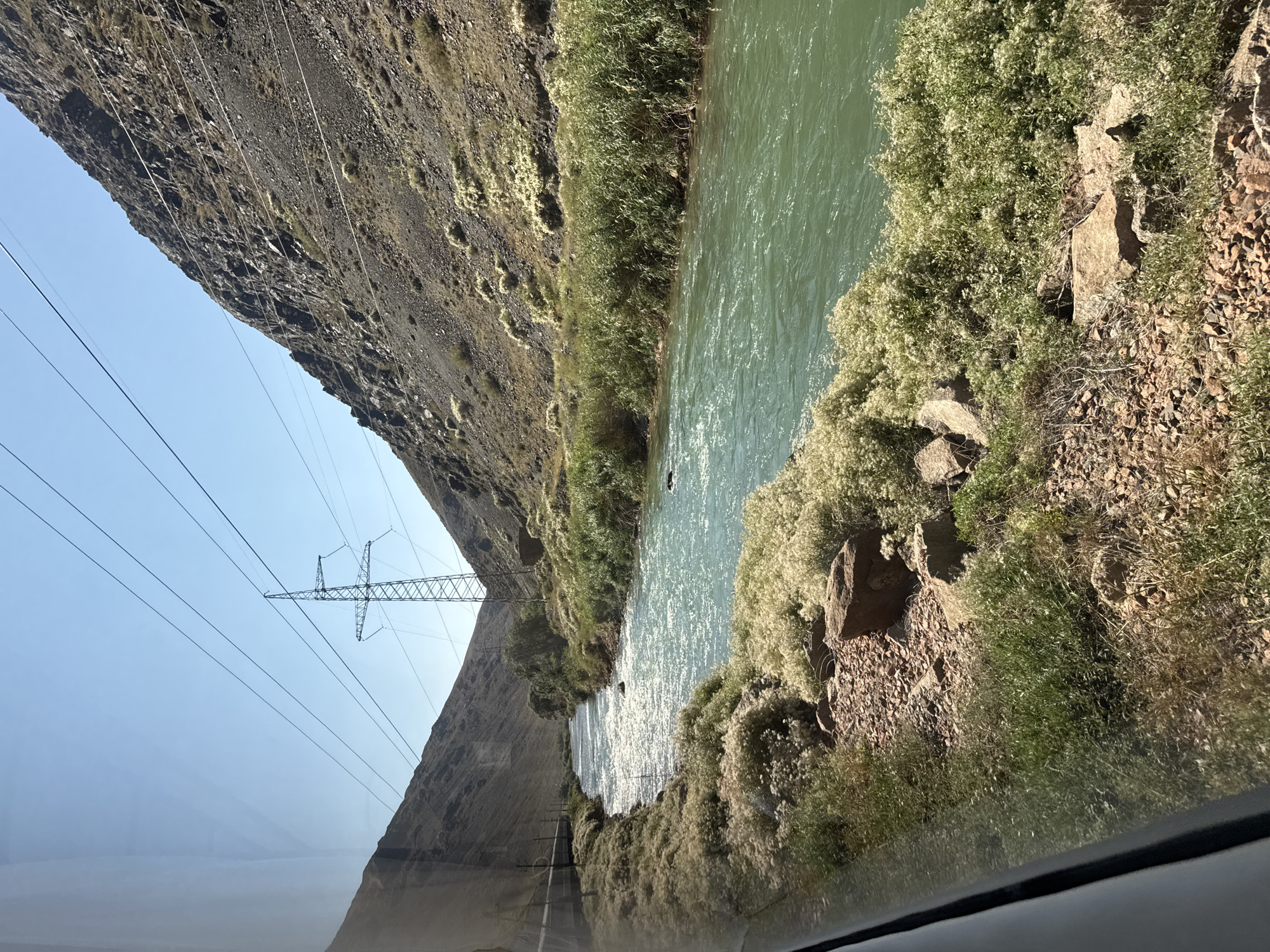 The image captures a wide, flowing river with turquoise-colored water cutting through a rugged, mountainous landscape. The riverbanks are lined with dense green reeds and interspersed with clusters of fluffy white flowering plants. The steep slopes of the mountains are covered in sparse vegetation and rocky terrain. Overhead, power lines stretch across the sky, supported by tall metal pylons, indicating human infrastructure in this otherwise natural setting. The sky is clear and blue, suggesting a sunny day. The perspective suggests the photo was taken from a moving vehicle, possibly a car or bus, as indicated by the faint reflection on the left side of the frame. The environment appears to be a remote, natural area characterized by a river, mountains, and vegetation, with the presence of power lines suggesting proximity to or a pathway through inhabited regions. The specific location within the city of Unknown, Kyrgyzstan, is not determinable from the visual elements present in the image.