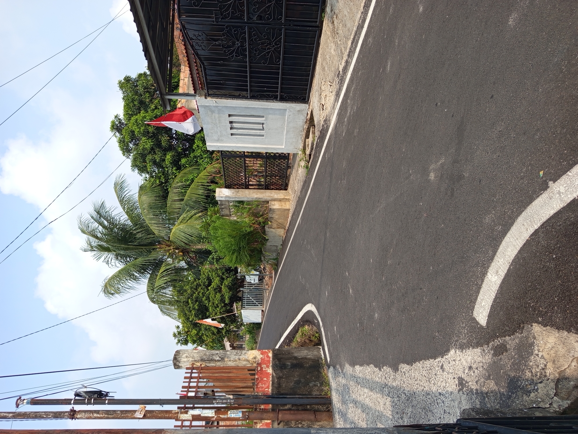 The image shows a narrow, paved road in Palembang, Indonesia, on a sunny day with a few clouds in the sky. The road curves to the left and appears to be in a residential or semi-rural area.
On the left side of the road, there is a tall, weathered utility pole with several wires running from it. Adjacent to the pole is a rusted metal fence and a concrete structure with red and white paint markings. A white stripe is painted on the road surface, indicating a lane divider or curve warning.
On the right side of the road, there is a large black metal gate with ornate scrollwork, suggesting it is the entrance to a property. Next to the gate is a light-colored concrete pillar with a rectangular window-like opening. A red and white Indonesian flag is flying from a pole near this pillar. Behind the gate and pillar, there is a red-tiled roof and more greenery.
Further up the road, more trees and houses are visible. A coconut palm tree is prominent in the middle ground, with its fronds spread out against the sky. The road surface is dark asphalt, appearing to be in good condition. The overall impression is of a quiet street scene in Palembang, possibly decorated for a national celebration due to the Indonesian flag. There are no people visible in the image.