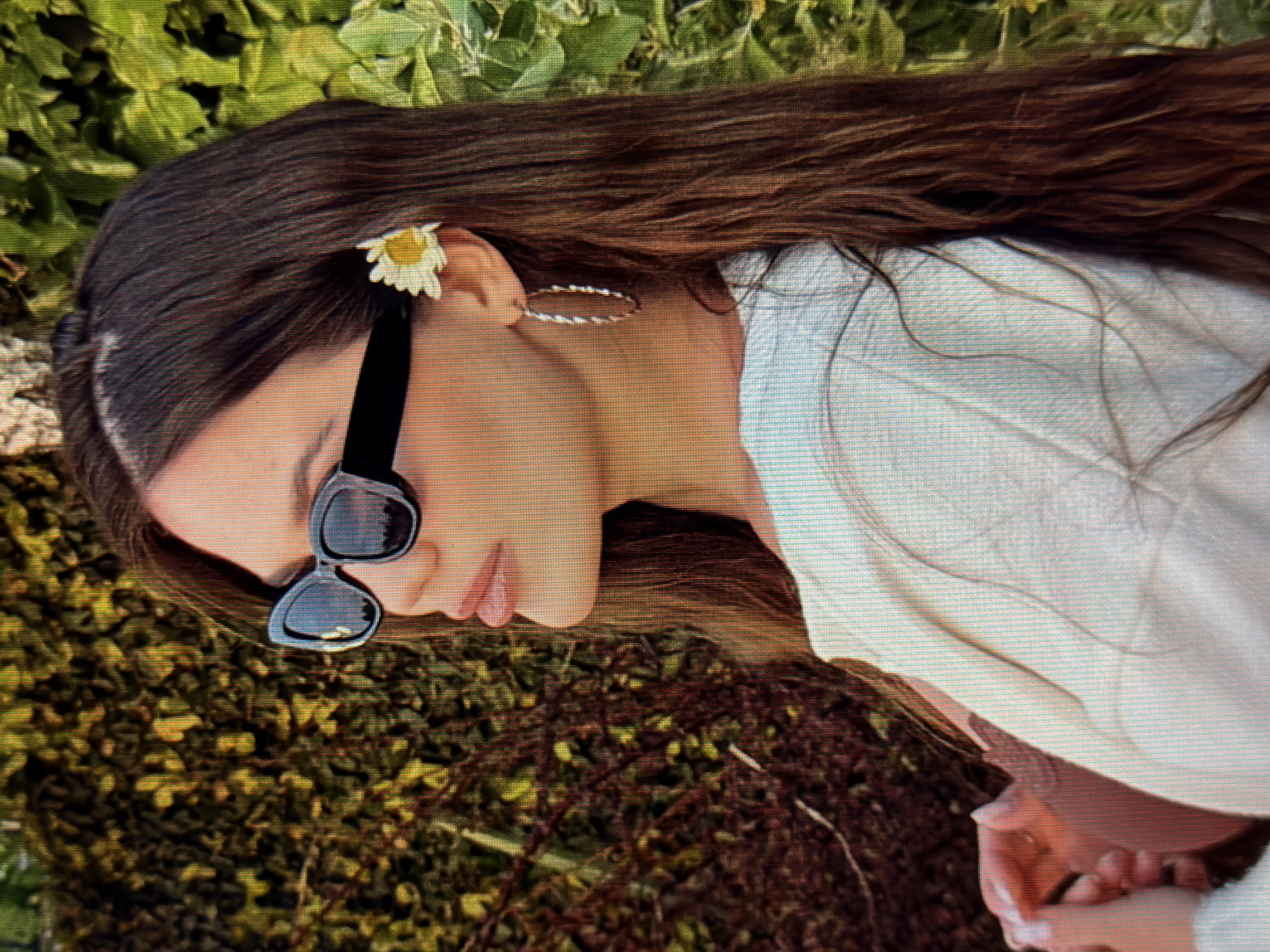 In the city of Sirius, Russia, a medium-shot profile view captures a young woman with long, dark brown hair, partially adorned with a single white daisy behind her right ear. She wears large, black, cat-eye sunglasses that obscure her eyes and her lips are slightly parted, revealing a subtle hint of her tongue. Her left ear is adorned with a silver, twisted hoop earring. She is dressed in a cream-colored, textured top or jacket. Her hands are clasped together in front of her chest, with her left hand visible resting on her right. The background consists of lush green foliage, with a hint of darker, reddish-brown branches or leaves on the left side, suggesting an outdoor, possibly garden or park setting. The lighting appears to be natural, casting a warm glow on the scene, likely indicating late afternoon or early evening sunlight.