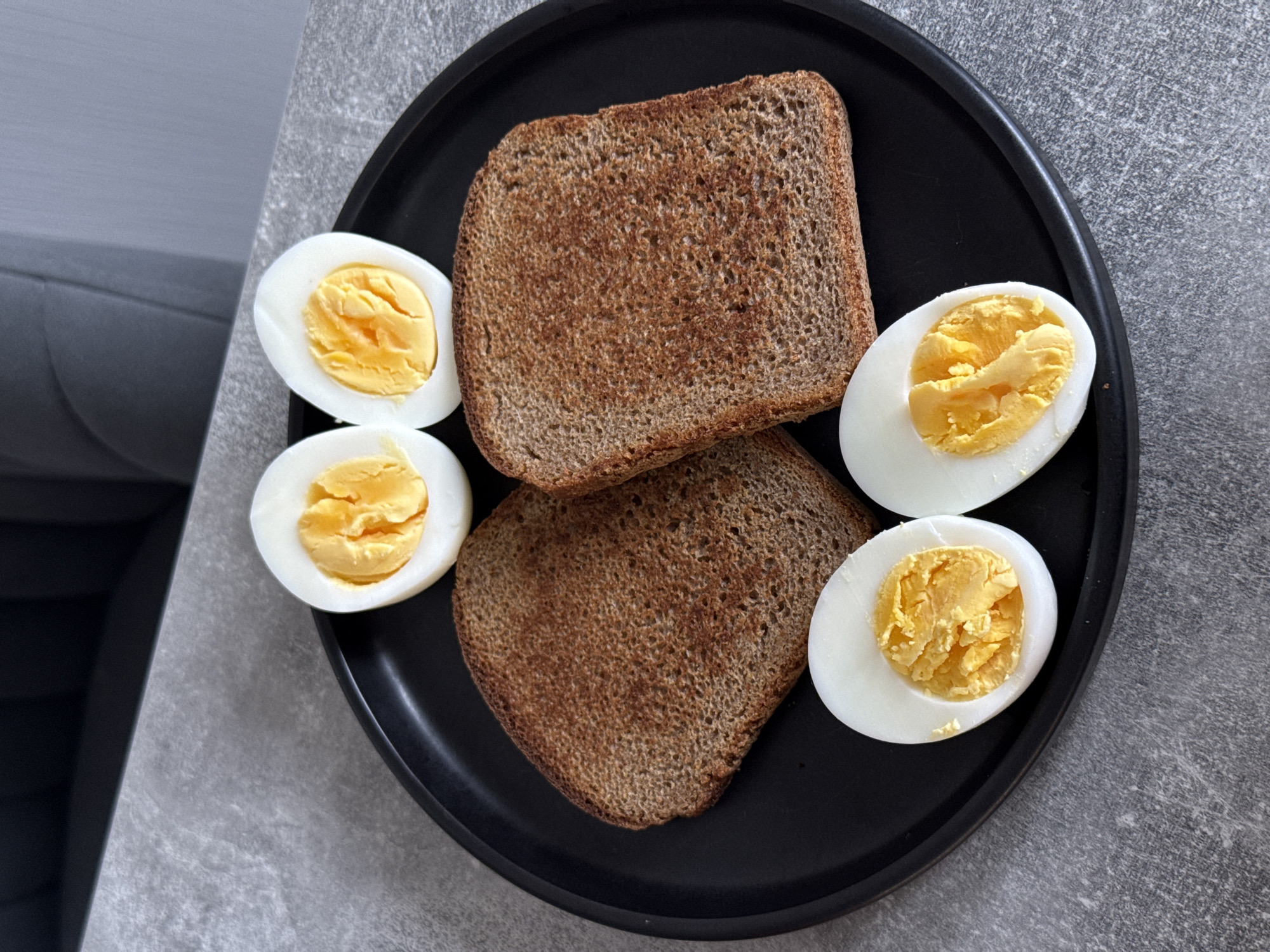 On a gray textured countertop in Kraskovo, Russia, a black plate holds a breakfast of two slices of dark toasted bread and four halves of hard-boiled eggs. The eggs are evenly cooked, with bright yellow yolks and white albumen. The toast is a deep brown, indicating it has been toasted for some time, and has a slightly uneven surface texture. The plate is positioned in the center of the frame, with the countertop extending to the edges of the image.