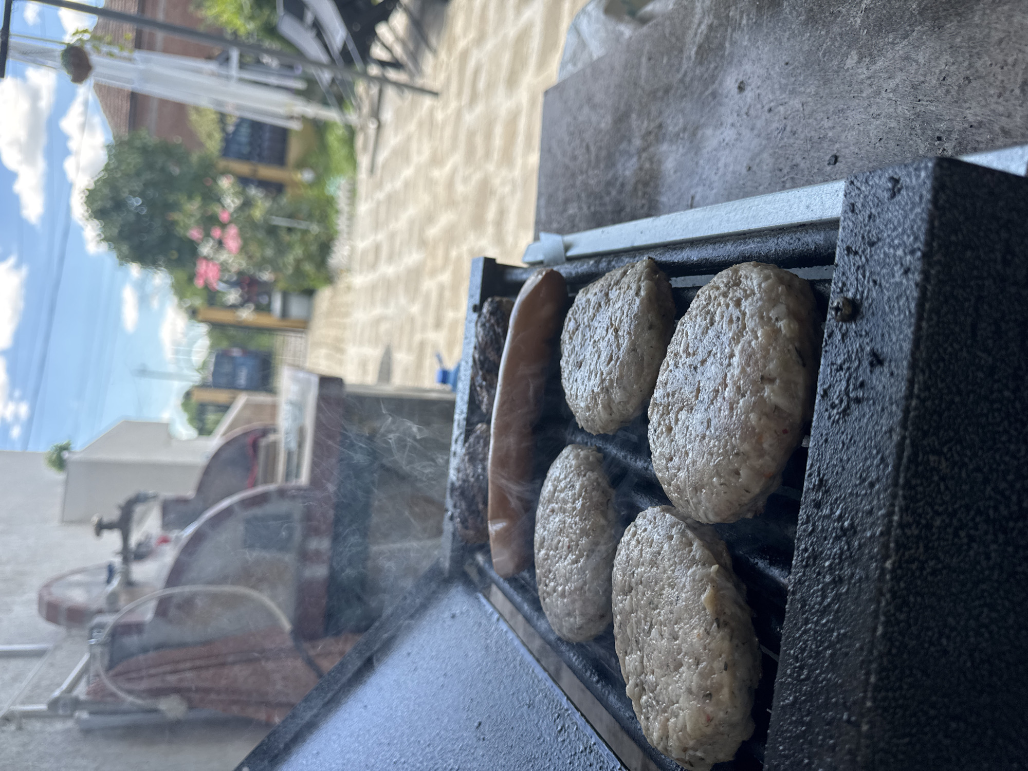 This is a close-up, eye-level shot of a grill with food cooking. In the foreground, four uncooked hamburger patties are arranged on the grill grates. Behind them, a single hot dog and two burger patties are also visible, with steam rising from the food. The grill itself appears to be a portable outdoor model. The background of the image shows a paved patio area, a wooden fence, lush green trees, and a blue sky with scattered clouds. The scene suggests an outdoor gathering or meal preparation, likely in a residential backyard. The image does not provide any information to locate it in the city of Rusokastro, Bulgaria.