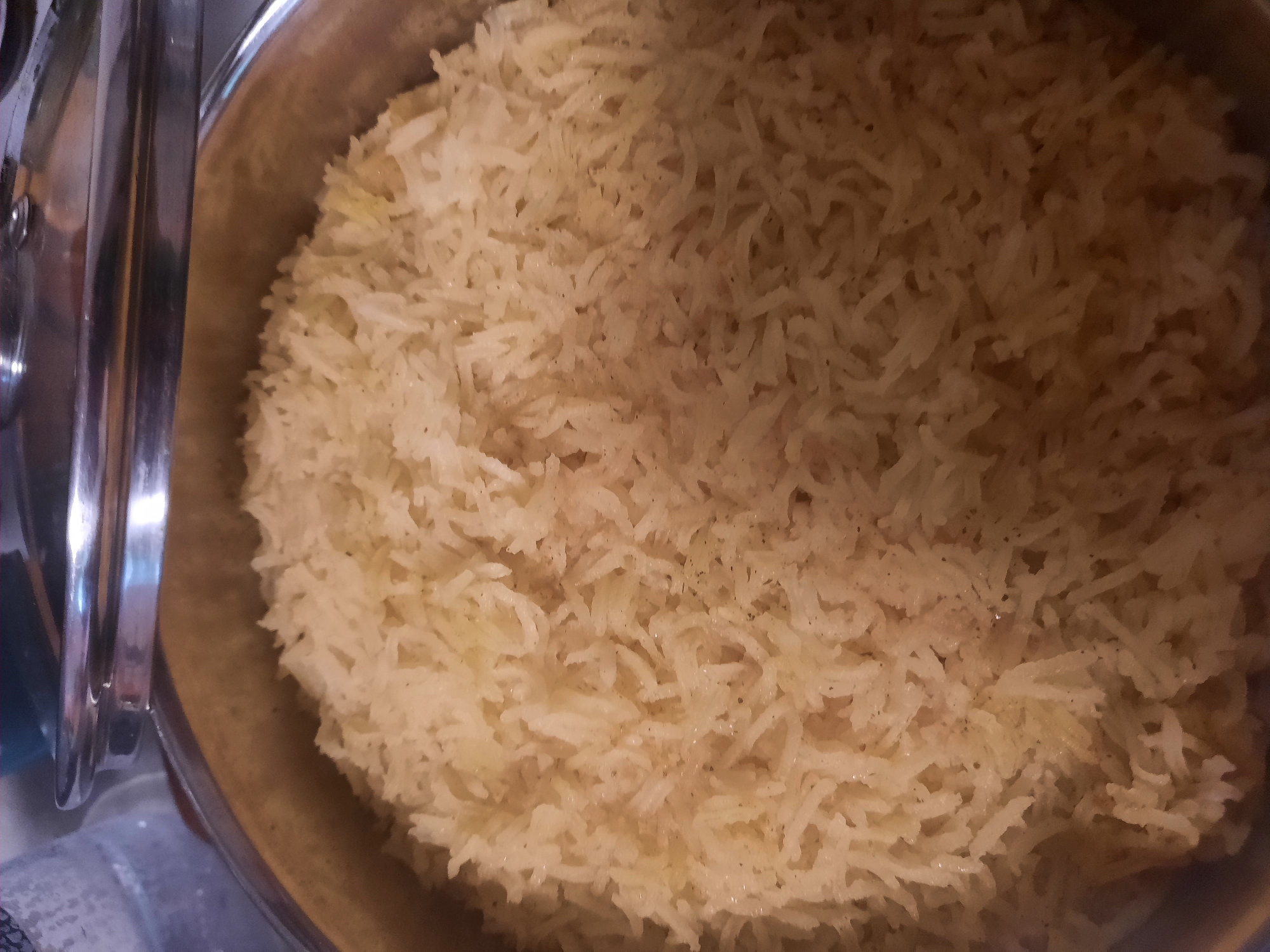 This is a close-up, overhead shot of a large quantity of cooked white rice, filling a metal pot or serving dish. The rice grains are long and slender, characteristic of basmati or jasmine rice, and appear fluffy and slightly separated, suggesting they have been prepared well. Some grains have very subtle darker specks, possibly indicating seasoning like pepper or herbs.
The setting appears to be indoors, likely a kitchen or dining area. To the left of the frame, a portion of a shiny, metallic serving utensil or part of a chafing dish lid is visible, with a reflective surface. The lighting is relatively soft and warm, suggesting ambient interior lighting, and does not provide strong shadows. There are no indications of time of day or weather.
The image focuses entirely on the food, with no people or clear environmental details apart from the serving vessel. The overall impression is one of abundance and preparation for a meal. No text is visible in the image. The implied location of Rosenheim, Germany, does not offer specific visual cues within this image of rice.