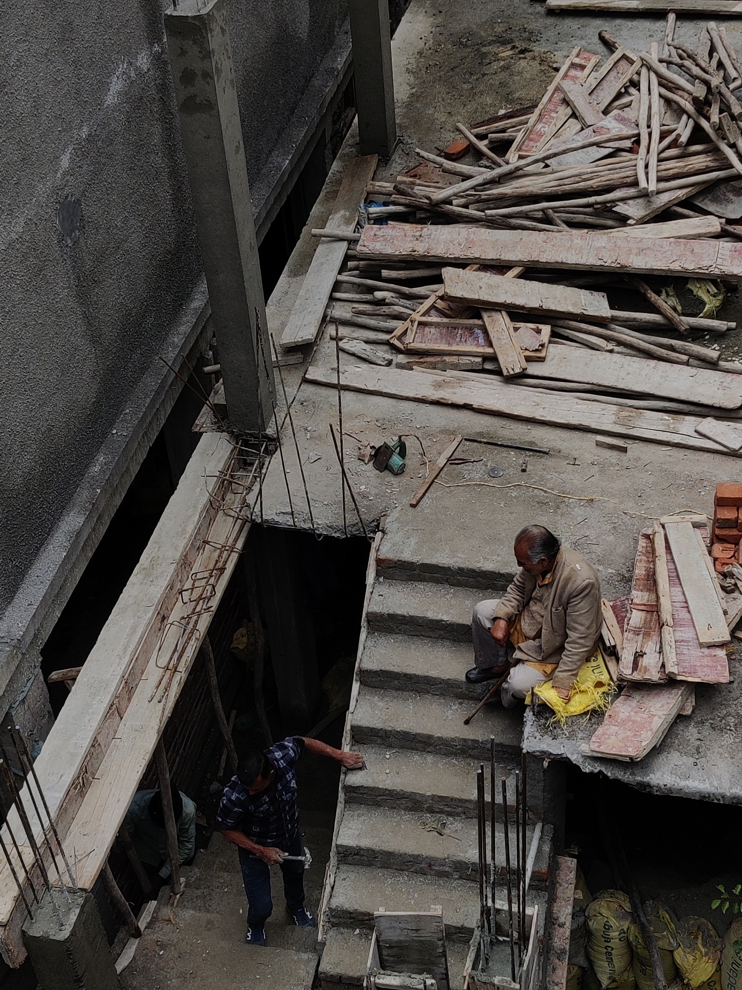 The image depicts an outdoor construction site in Vashisht, India, with a focus on unfinished concrete structures, wooden scaffolding, and materials. In the foreground, a man in a light brown jacket and grey pants sits on a concrete staircase, looking down, with a yellow bag near his feet. To his left and lower down the stairs, another man in a blue and black checkered shirt and dark pants is working, holding a hammer and seemingly hammering something. A third person in a green shirt is visible further down in a darker area. The environment is characterized by rough, grey concrete surfaces, stacks of wooden planks and poles, reinforcing steel bars protruding from concrete, and several large yellow sacks, possibly containing cement. The overall scene suggests active construction work in progress.