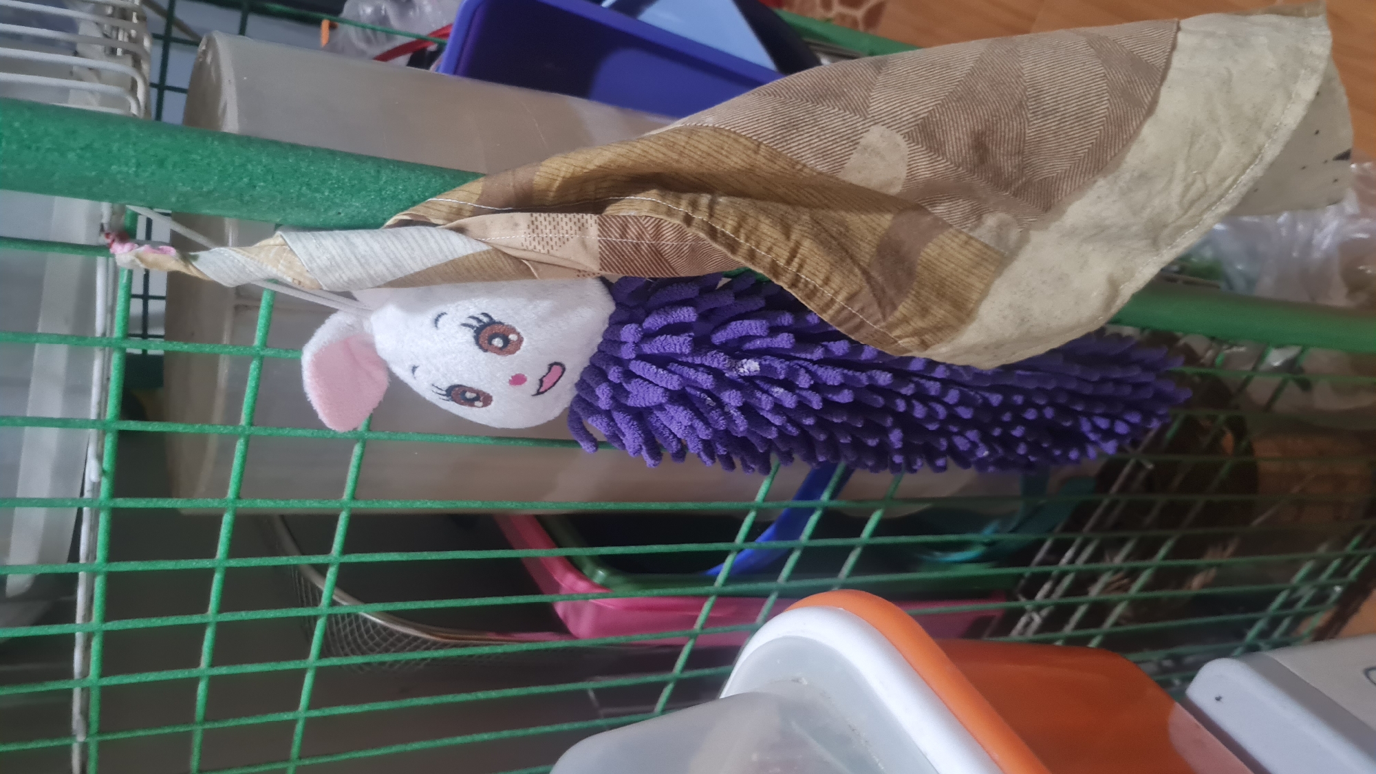 In Bandar Lampung, Indonesia, a plush white rabbit toy with pink ears and embroidered facial features hangs from a green metal shelf unit. The toy has a purple, fluffy body that resembles a duster. A tan and brown patterned cloth is draped over the rabbit and hangs down. Behind the rabbit and cloth, various household items are visible, including plastic containers and what appears to be a metal bucket. The environment suggests a storage or utility area within a home.