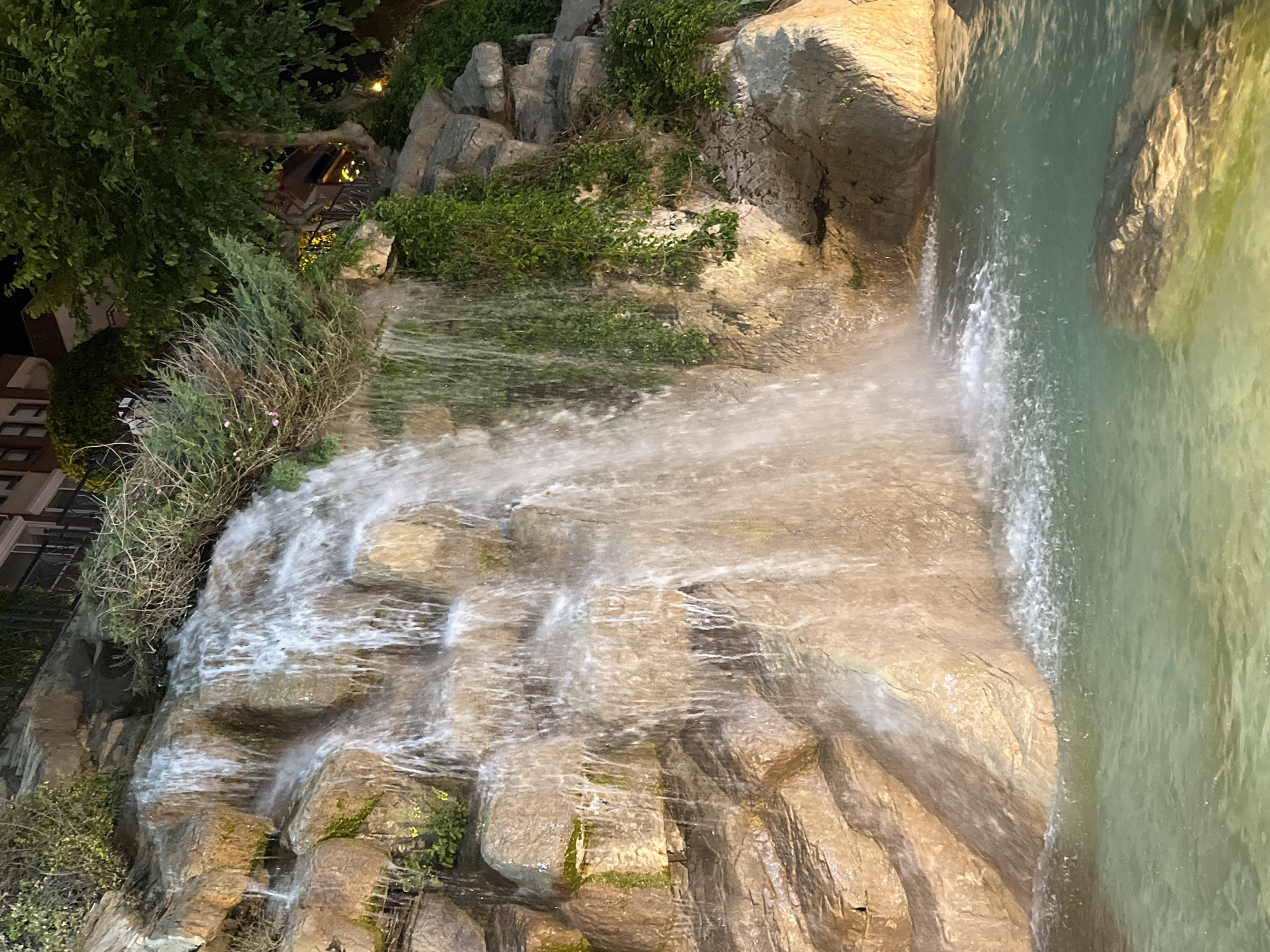 This image captures a beautifully illuminated waterfall feature at night in Alanya, Turkey. The central focus is a multi-tiered cascade of water flowing over rough, light-brown and grey rocks. The water appears frothy and white as it tumbles downwards, eventually collecting in a calm, greenish-blue pool on the right side of the frame.
Lush green vegetation, including small bushes, grasses, and climbing vines, adorns the rock formations and borders the waterfall, adding to a natural aesthetic. On the far left, larger, darker trees and foliage are visible, suggesting a more extensive garden or landscape. The scene is lit by artificial lights, casting warm glows on the rocks and the moving water, which indicates the image was taken during the evening or nighttime. Distant warm-toned lights can be vaguely seen through the upper left foliage, hinting at nearby buildings or decorative lighting.
There are no people or visible text in the image. The overall impression is one of tranquility and natural beauty, highlighting the peaceful flow of water in a well-lit, serene setting.