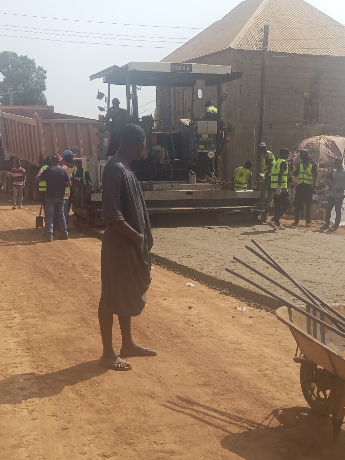 The image depicts an active road construction scene on a dirt road in Akwanga, Nigeria, under a bright, hazy sky. In the left foreground, a dark-skinned male, wearing a dark gray tunic, baggy shorts, and flip-flops, stands facing right with his hands in his pockets, observing the activity. To the right of the foreground figure, a metal wheelbarrow containing several metal rods is positioned on the reddish-brown dirt ground.
In the midground, a large asphalt paver, branded "Implenia," is actively laying a dark, new road surface. A large orange dump truck with its bed raised is positioned to the left of the paver, appearing to offload material into it. Multiple construction workers are present around the machinery; several are wearing neon yellow high-visibility vests and blue caps, engaged in the paving operations. One worker is visible operating the paver.
The background features a two-story building with light-colored concrete block walls and a prominent pyramid-shaped, light brown corrugated metal roof. Utility poles and overhead wires are visible against the bright sky. The ground transitions from a reddish-brown dirt road, showing scattered debris, to the newly paved asphalt section. Shadows are cast towards the left, indicating a light source from the right.