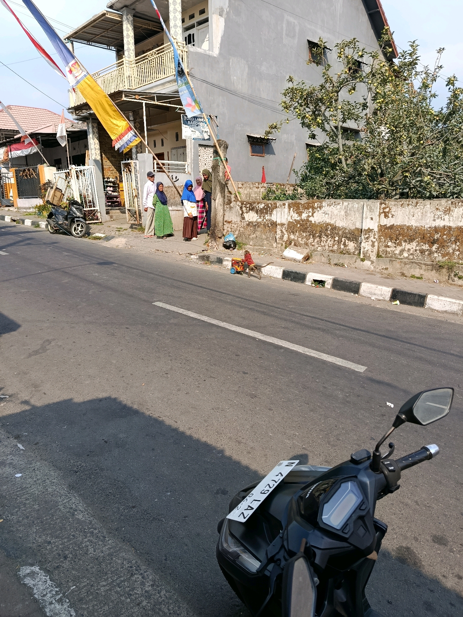 The image depicts a street scene in Gubukklakah, Indonesia, with a focus on a residential area bordering a road. In the foreground, a black motorcycle is parked, its handlebars and a portion of its front panel visible. The road surface is asphalt with a white lane divider. To the left of the road, a sidewalk is present, with a dark-colored motorcycle parked on it. Several people, including two women in hijabs and one man, are standing on the sidewalk near the entrance to a building. The building is a multi-story structure with balconies and a grey facade, adorned with flags and banners. A tree with visible fruit is growing near the building. Further down the sidewalk, a small monkey is seen walking near a small cart with wheels. The environment is characterized by residential buildings, a street, and vegetation. The time of day appears to be late afternoon, judging by the shadows.