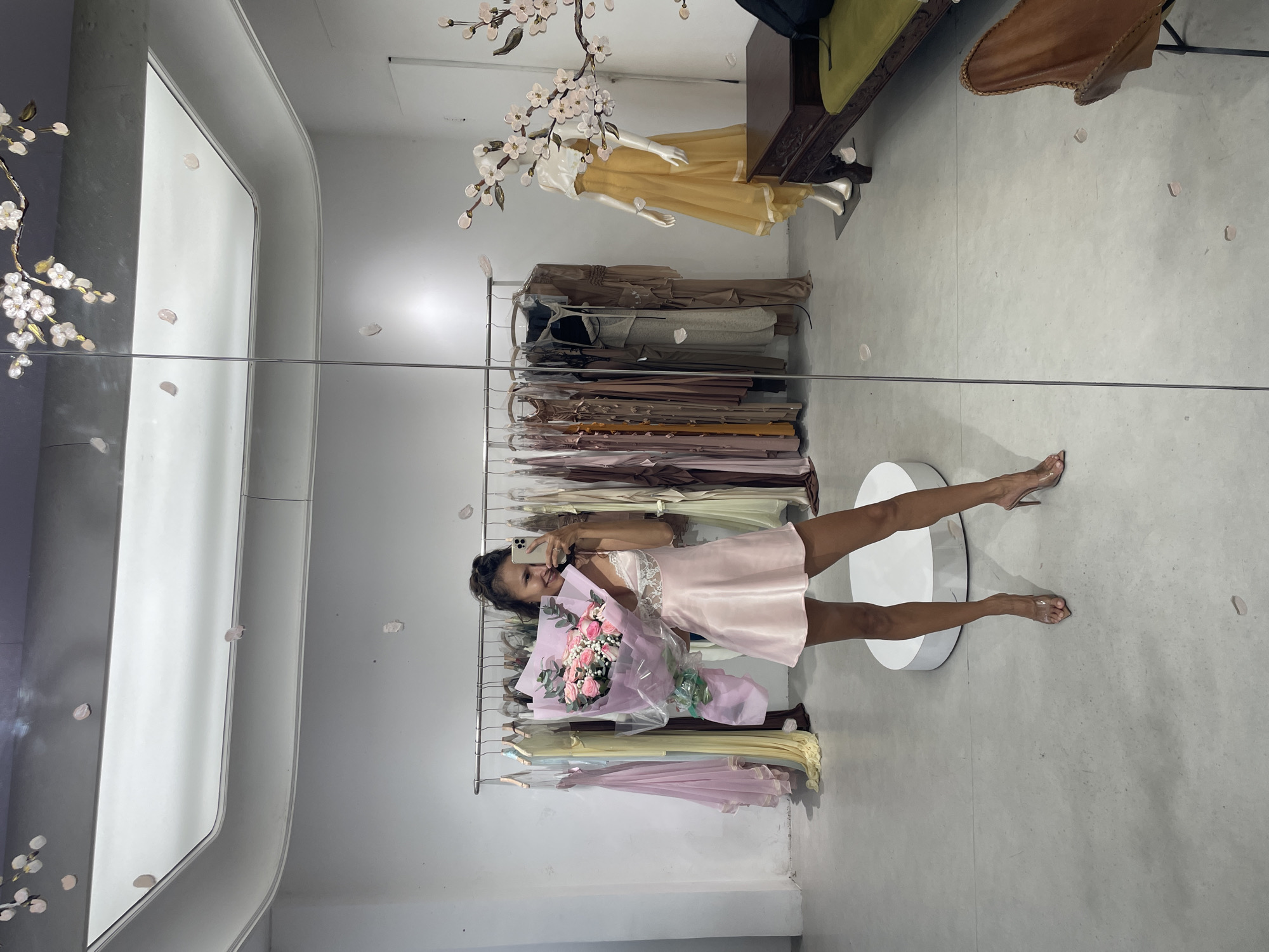 The image displays the interior of a clothing boutique in Thủ Đức, Vietnam. A woman is captured in the reflection of a large mirror, smiling and holding a substantial bouquet of approximately two dozen pink roses, wrapped in light purple paper and clear plastic with a green ribbon. She is wearing a light pink, satin-like slip dress featuring white lace on the bodice, and clear high-heeled mules. Her dark hair is styled up, and she is holding a light-colored smartphone to take the photograph.
The background, visible through the mirror, consists of white walls and a light gray concrete floor. Two clothing racks extend across the scene, laden with numerous garments, primarily dresses and flowing pieces, in pastel shades of pink, yellow, brown, and cream. To the far right, a mannequin is dressed in a flowing yellow gown with a white top. A decorative branch adorned with pink and white cherry blossom-like flowers is attached to the wall/mirror frame in the upper right quadrant. Light pink petals are scattered across the floor and reflected on the white, recessed-lit ceiling. Additional furnishings include a dark wooden bench with a green cushion, a black backpack resting on the bench, a light brown leather butterfly chair in the bottom right corner, and a round white display pedestal on the floor.