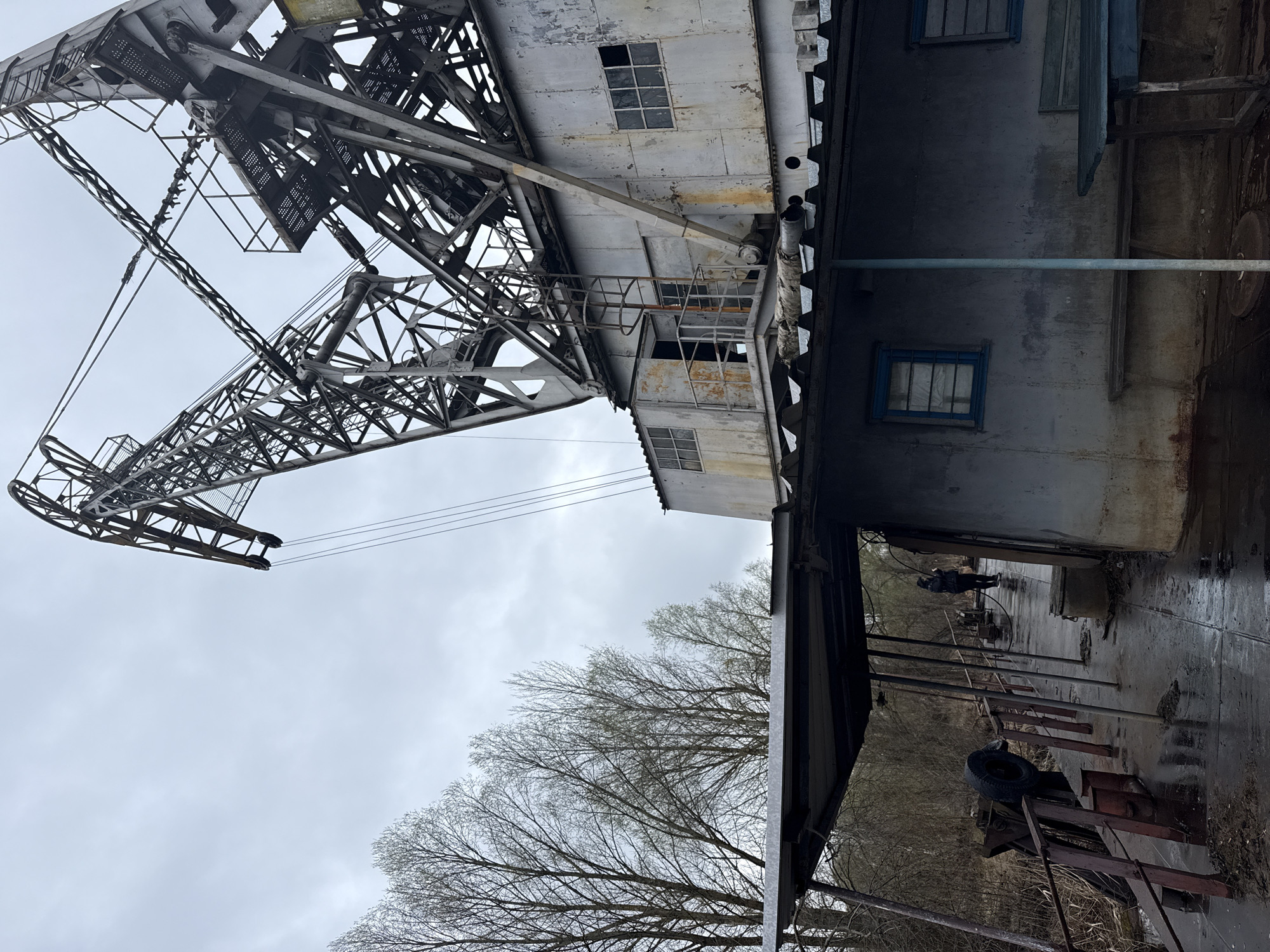 The image shows a large, industrial crane against a grey sky and bare trees. The crane appears metallic and aged. Attached to the crane is a weathered, multi-story structure with windows. The base of the structure extends horizontally and features rusted metal equipment. A solitary figure is walking. This structure appears to be in the city of Chornobyl, Ukraine.
