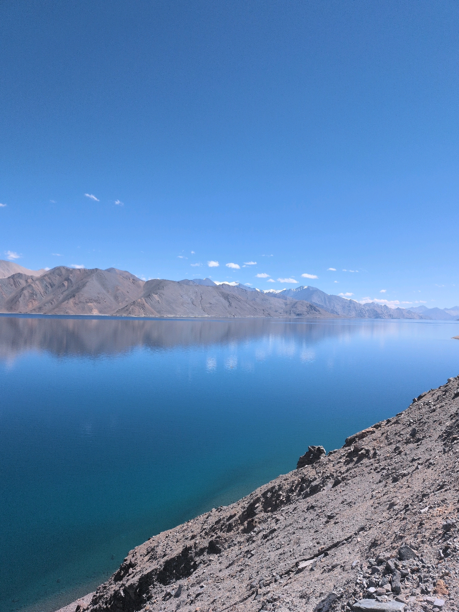 The image depicts a serene high-altitude lake with a clear blue sky overhead. The water of the lake is a vibrant, deep blue, reflecting the surrounding mountains and the sky. The mountains, characterized by their rugged, rocky terrain and muted gray and brown hues, form a long, undulating silhouette across the horizon. Some of the peaks in the distance appear to be capped with snow. The foreground of the image features a steep, rocky incline covered with loose stones and gravel, sloping down towards the lake. The overall scene conveys a sense of vastness and natural beauty, typical of a mountainous, remote landscape. The location is identified as Pangong Tso, in Ladakh, India, a transboundary lake that lies on the border between India and China.