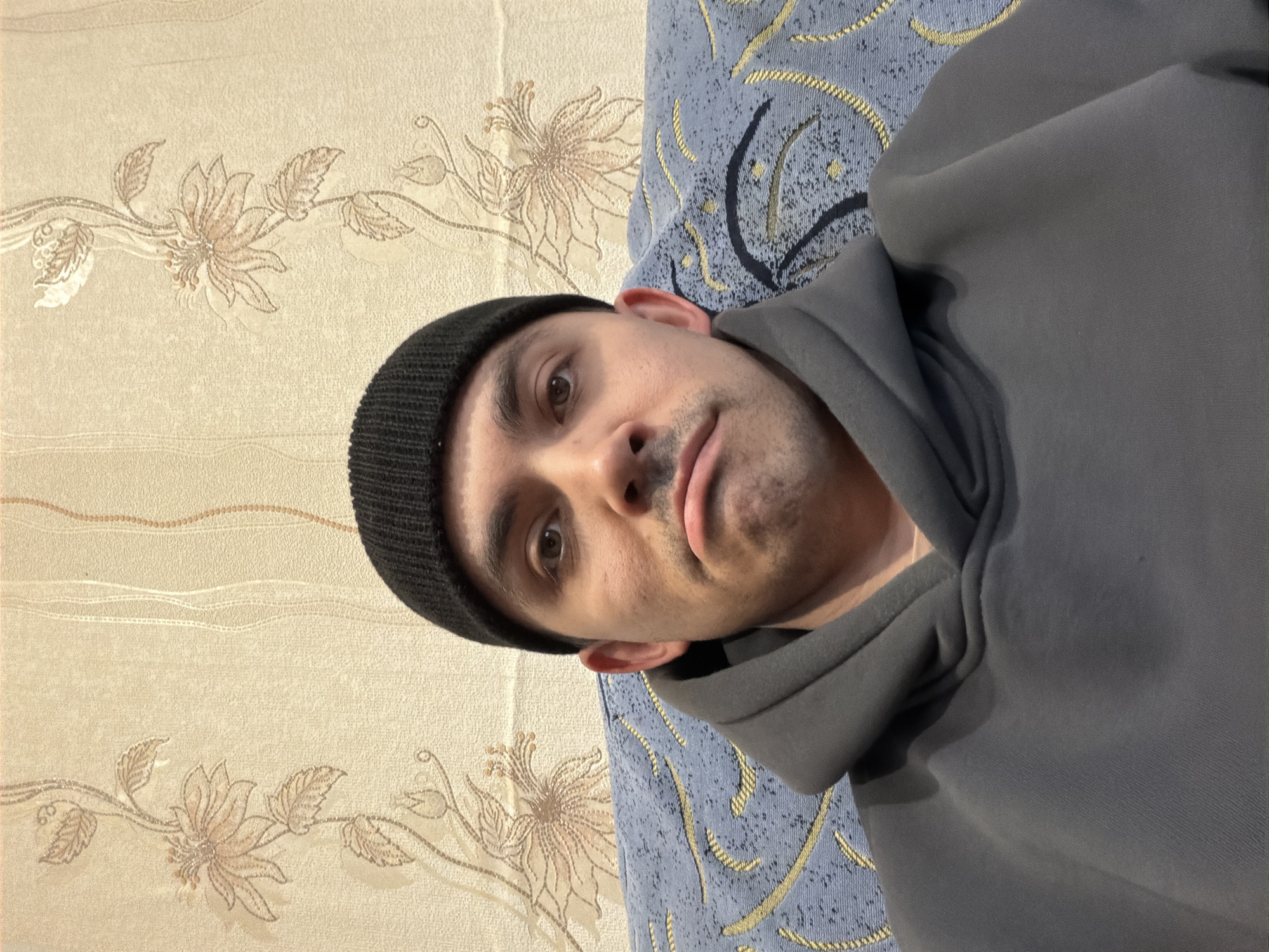 A close-up, rotated portrait photograph shows a man wearing a black beanie and a gray hooded sweatshirt. He is lying down, with part of a blue patterned sofa visible behind him and wallpaper with a floral pattern to his left. The man has brown eyes, dark hair, and stubble. He is looking directly at the camera with a neutral expression. No specific information indicates the location is Izhevsk, Russia; this detail is not discernible from the image.