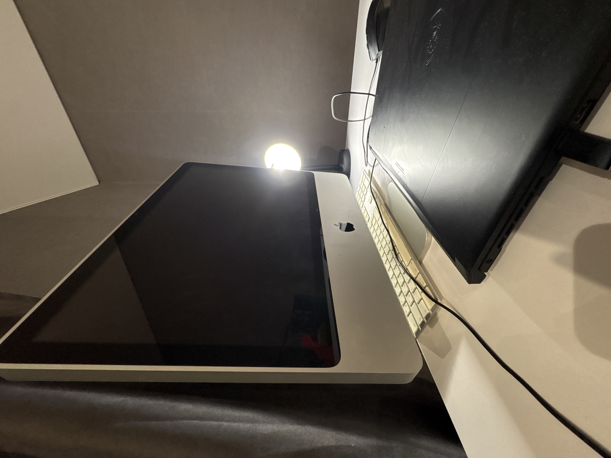 The image shows a desk setup. There is a large tablet computer with a black screen, likely turned off. A keyboard is present next to it, and a second, larger monitor is positioned perpendicular to the tablet. A desk lamp illuminates the scene from above. The environment is a room with gray walls. The location and city are not identifiable from the image.
