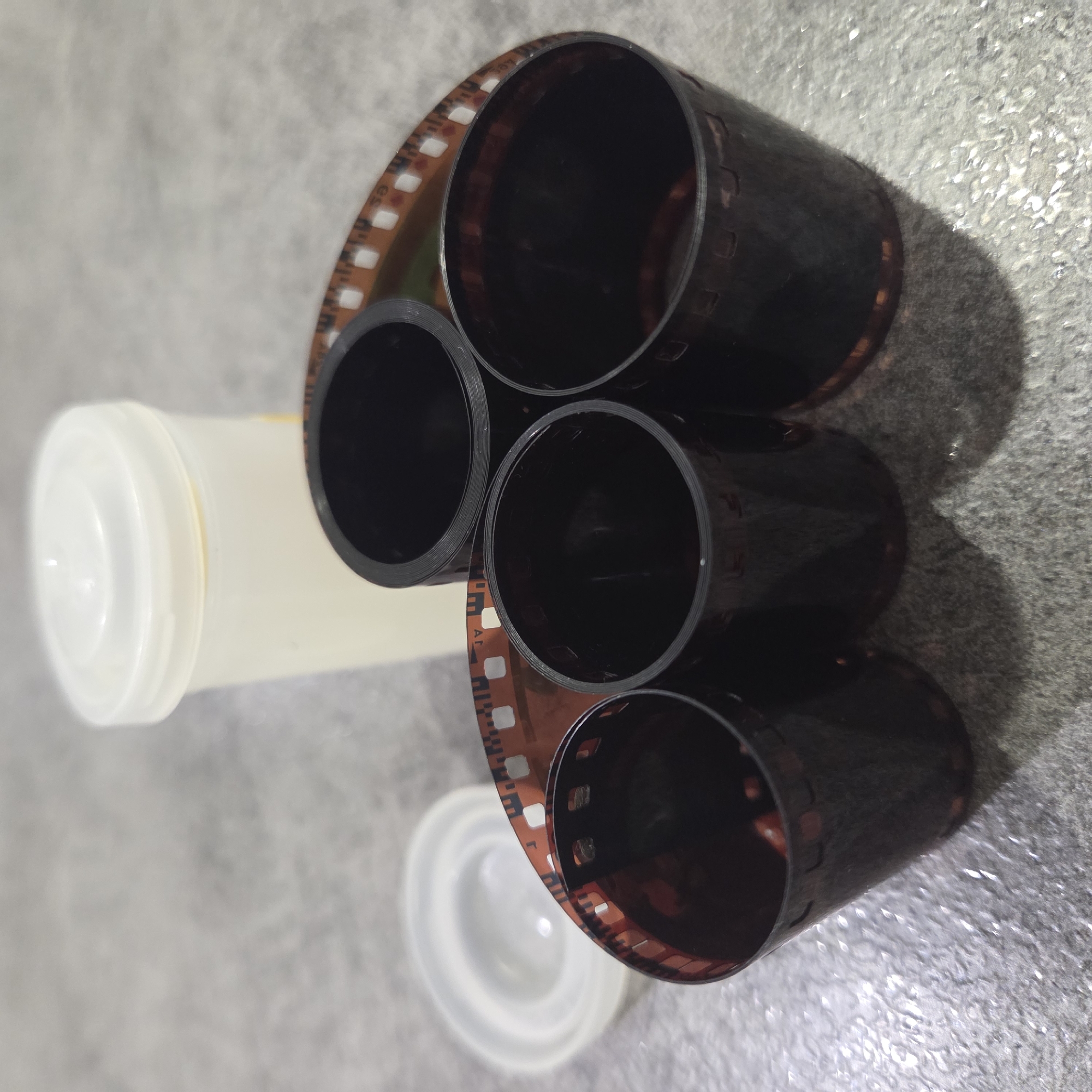 The image presents a close-up, top-down view of several rolls of 35mm photographic film laid out on a textured, light gray surface. Four main rolls of film are prominently displayed, appearing as dark, hollow cylinders due to the tightly wound film strips. The film itself features a brownish-orange base and characteristic sprocket holes running along both edges. The dark emulsion side of the film is mostly facing inwards, typical of processed or unexposed film within a roll.
A light-colored, translucent plastic film canister with a white lid is partially visible on the left side of the frame, positioned horizontally. Another white lid, likely from a similar canister, is seen in the lower right corner. The film rolls are arranged somewhat informally, with some overlapping, suggesting they are either in storage, have been recently used, or are being organized.
The background is a mottled, textured gray surface, possibly a tabletop or floor with a concrete-like finish, exhibiting some reflective highlights indicating its uneven nature. Shadows cast by the film rolls suggest an overhead or angled light source, consistent with an indoor setting. Faint text and numbers, including what appears to be "AR", "es", and digits like "10" or "0", are visible along the perforated edges of the film, which are typical manufacturer and frame markings found on 35mm film. No people are present, and no specific activity or event is taking place beyond the static arrangement of the film and canisters. The scene conveys no particular emotions.