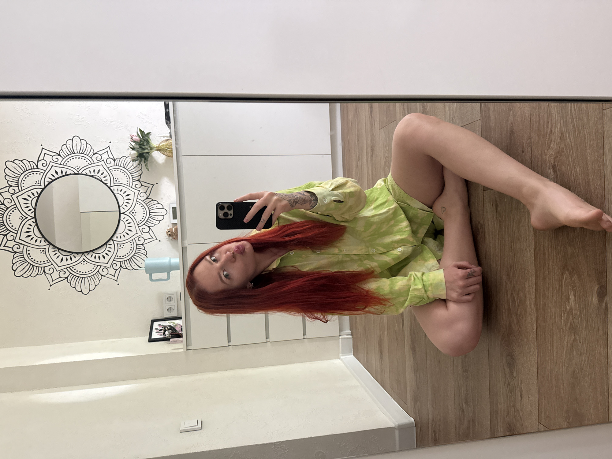 A woman with long, red hair is sitting on a wooden floor, taking a selfie in a mirror. She is wearing a light green tie-dye pajama set. In the background, there is a white dresser with some decorative items on top, including a mandala decal on the wall above it, a vase with flowers, and a framed picture. The room appears to be a bedroom or living space. There are no identifiable landmarks or contextual clues in the image to suggest the specific city of Rostov-on-Don, Russia.