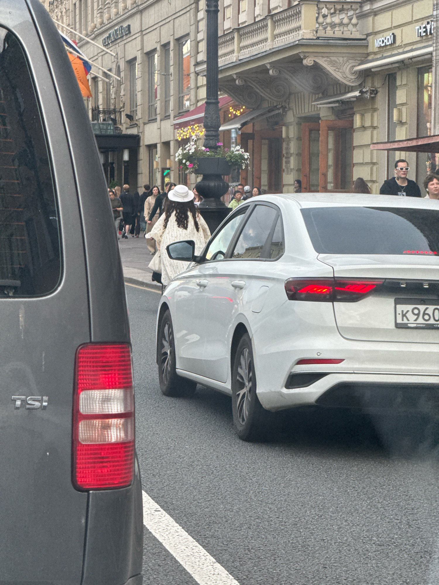 The image captures a street scene in Saint Petersburg, Russia, with traffic and pedestrians on a slightly overcast day. In the foreground, the rear of a dark grey vehicle is visible on the left, showing its taillight and the "TS!" emblem. To the right, a white sedan is in motion, displaying its rear, including its taillights and a license plate reading "K960". A person wearing a white hat and a patterned coat is walking in the middle ground, ahead of the white sedan.
The background features a European-style building with ornate architecture, balconies, and arched doorways. A street lamp with flower pots is also present. People are seen walking on the sidewalk, some appearing to be tourists. Signs on the building include "МЯСНОИ" and "THE VE". The overall impression is of a busy urban environment in Saint Petersburg.