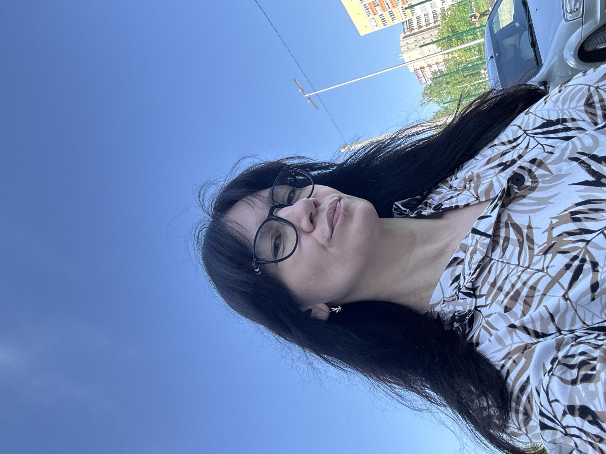 A medium-angle, rotated outdoor shot depicts a woman with dark hair and glasses, wearing a light-colored patterned blouse. She is looking at the camera with slightly pursed lips. In the background, there is a clear blue sky. In the upper part of the image, there are buildings, trees, a streetlight, and a portion of a car visible. Without further information, determining the specific location in Chaikovsky, Russia is not possible.