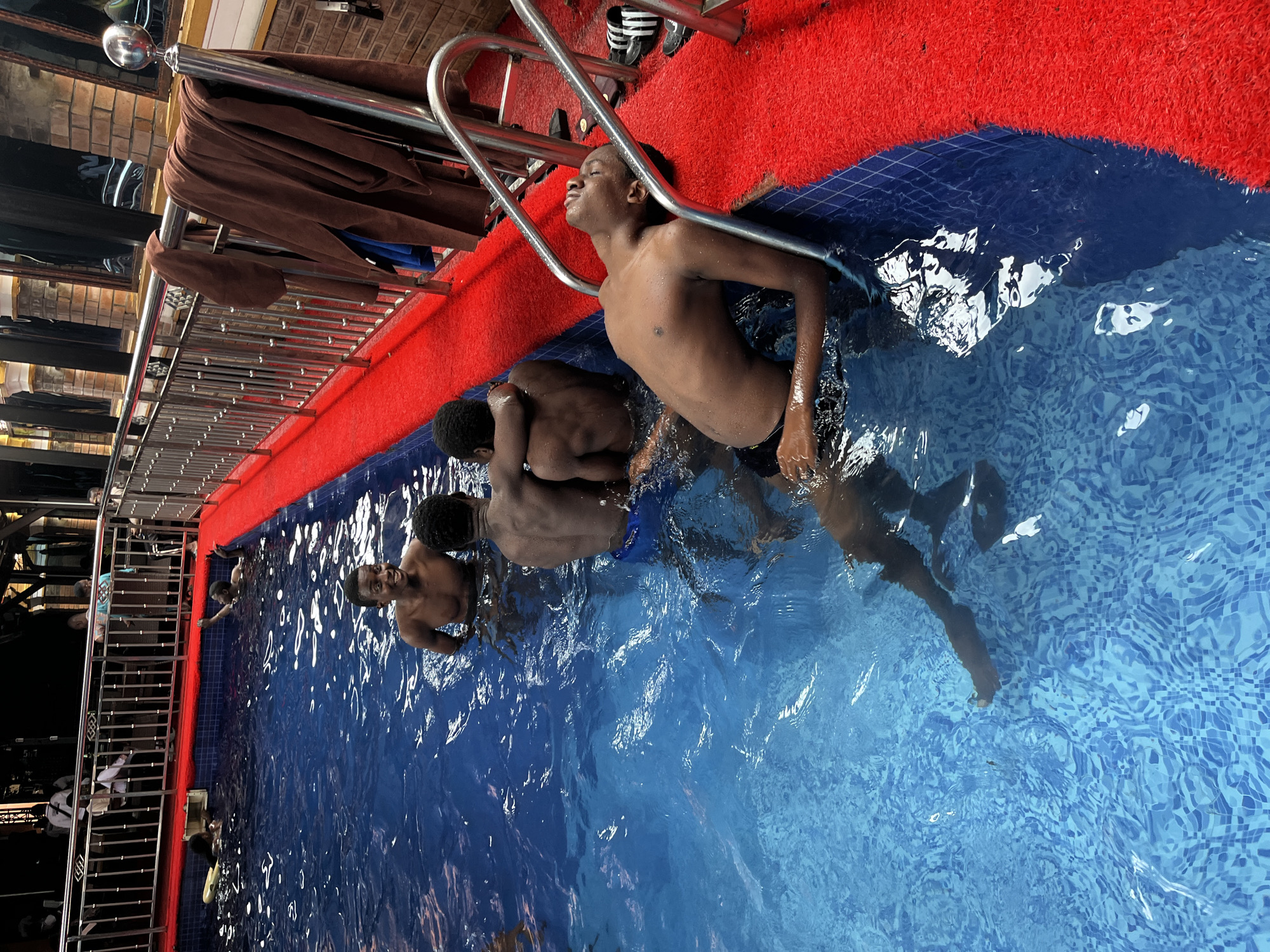 In Obosi, Nigeria, at a swimming pool, five young males are present. One individual, with closed eyes and head tilted back, leans against the pool ladder. Another male, with a wide smile, is visible from the chest up in the foreground, looking towards the left. Behind him, three other males are in the water, two of whom are embracing. The pool has blue tiled walls and is surrounded by a red carpeted deck. Metal railings are installed around the perimeter of the pool. Towels are draped over the railings.