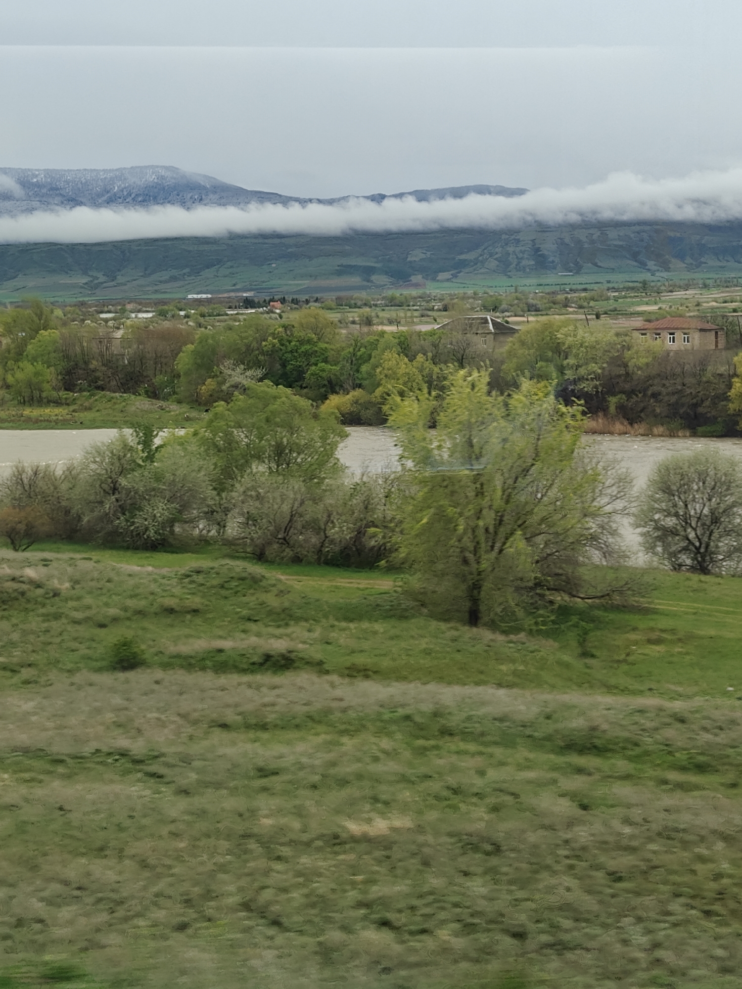 The image shows a landscape scene with a river flowing through a valley. The river is a muddy brown color, and there are trees and bushes growing along its banks. In the background, there are hills or mountains with snow-capped peaks partially obscured by clouds. The foreground is a grassy field with some small hills. Several buildings are visible in the background, indicating a settlement. The sky is overcast and grey.