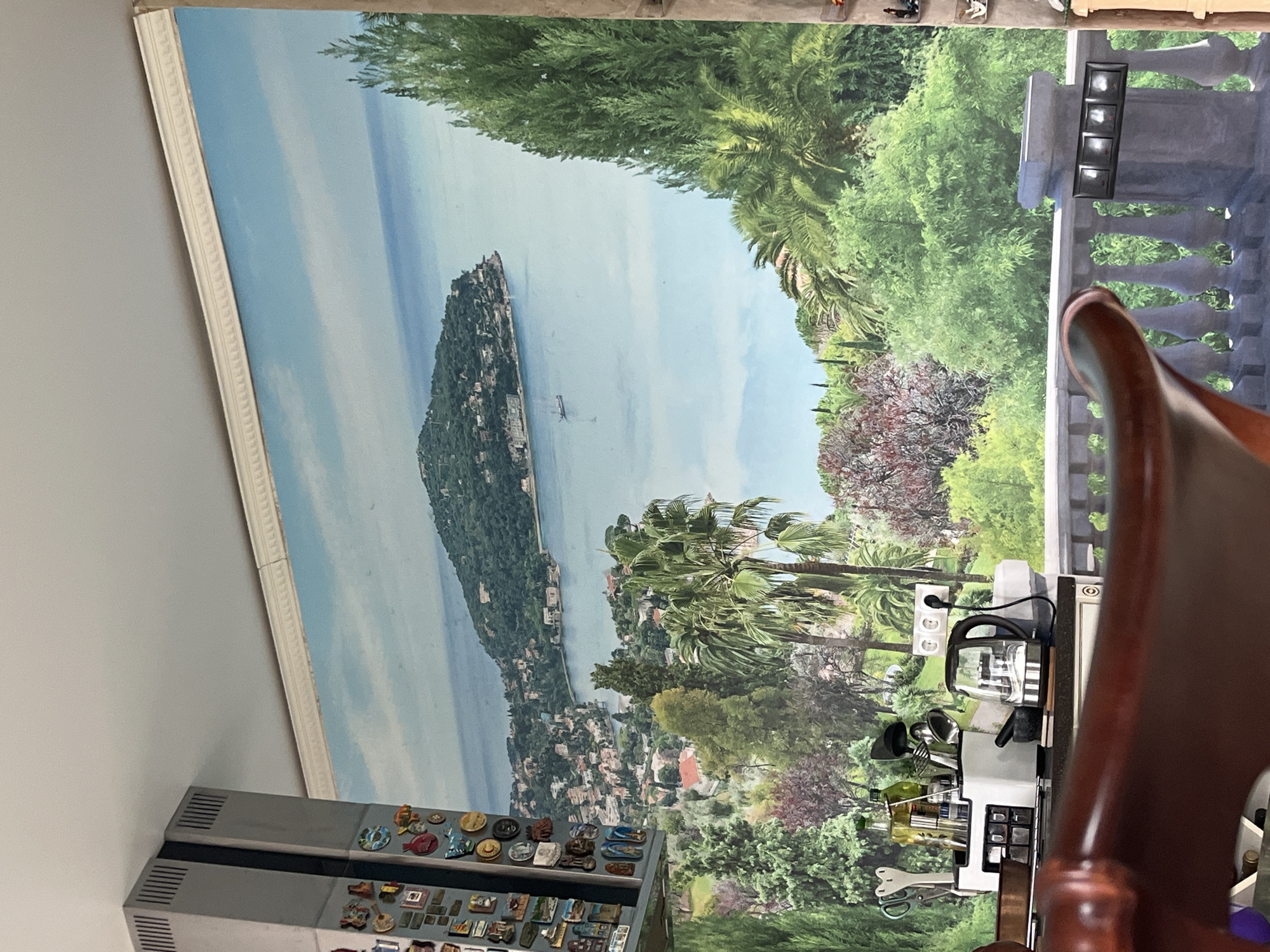 The image depicts an indoor scene with a large mural covering the back wall, creating the illusion of a Mediterranean landscape with a body of water, an island, and lush greenery. A refrigerator with numerous magnets on its side is visible to the left. In the foreground, a dark wooden chair back is partially visible, and a countertop with kitchen appliances, including a kettle and a mortar and pestle, is present. A decorative railing runs across the bottom of the frame, in front of the mural. The provided crops highlight different sections of the image, showing details of the mural's landscape, the refrigerator's magnets, and the kitchen counter. There is no information in the image or the provided crops that indicates the location is Vladimirovka, Russia.