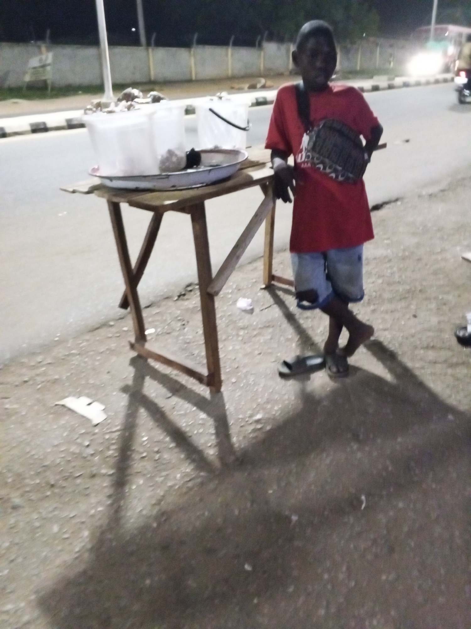 A young boy stands next to a makeshift wooden table on the side of a road in Adankolo, Nigeria. He is wearing a red t-shirt, denim shorts, and sandals, with a patterned crossbody bag. On the table are two white buckets, one partially obscured, and a metal tray. A streetlight illuminates the scene, casting long shadows. In the background, a concrete barrier lines the road, with traffic visible further in the distance. The ground is paved with asphalt. Debris is scattered near the table's base.