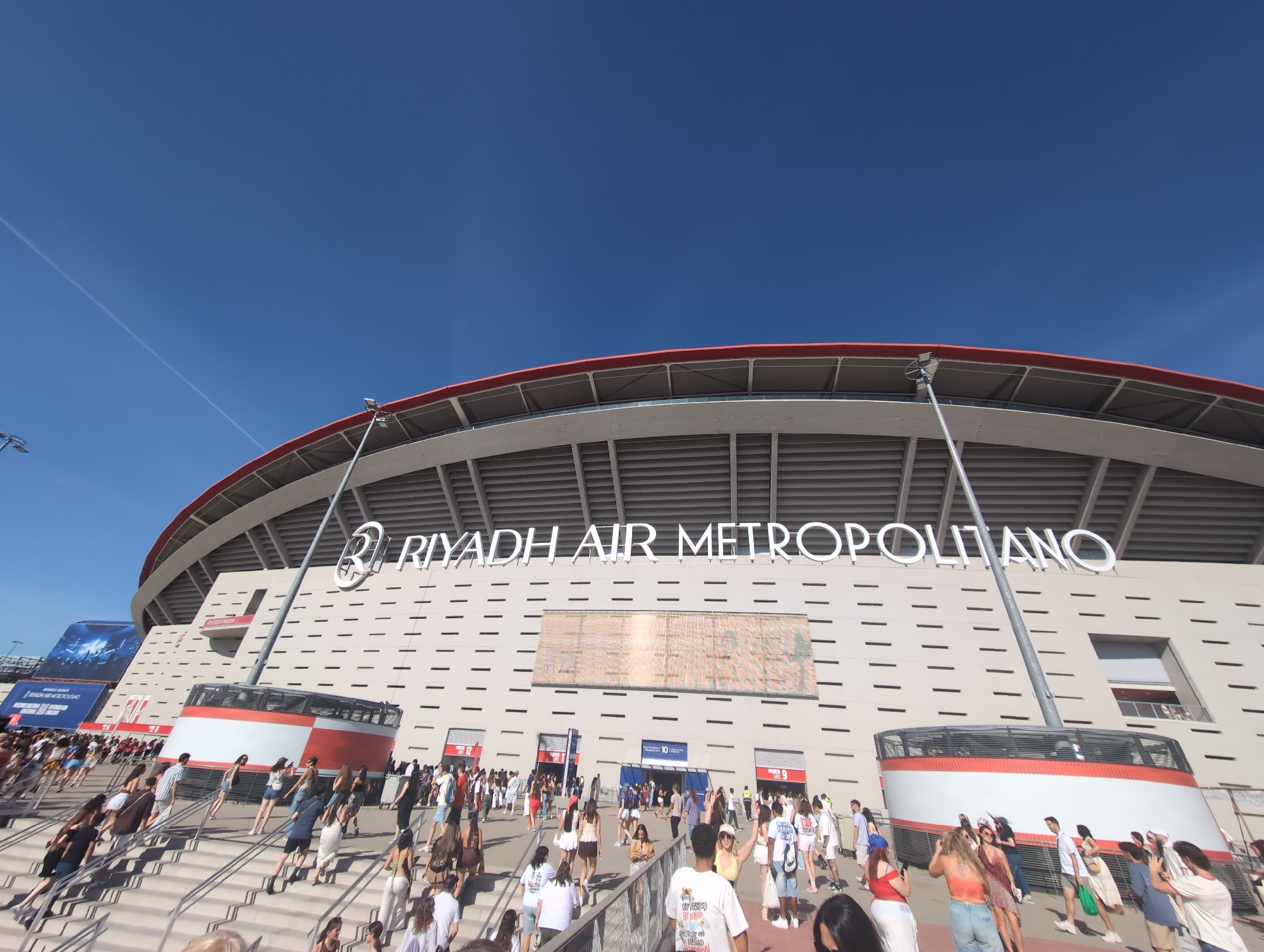 A wide-angle exterior shot captures the front of the Civitas Metropolitano stadium, home to Atlético Madrid, on a clear, sunny day. The stadium's curved roof, a prominent red structure, is visible against a bright blue sky with faint contrails from an aircraft. The stadium's facade is predominantly light-colored with numerous horizontal indentations, and large white lettering spells out "RIYADH AIR METROPOLITANO" across the top section. Several people are seen walking up a wide set of outdoor stairs leading to the stadium entrances, suggesting an event is about to begin or has just concluded. Some individuals are dressed casually in t-shirts, shorts, and dresses, while others appear to be carrying bags. Prominent banners and signage are mounted on the stadium's exterior, including a large digital screen displaying what appears to be event information. Red and white cylindrical structures, possibly part of the stadium's ventilation or artistic design, are positioned near the base of the building. The overall scene depicts a busy public gathering at a major sports venue.