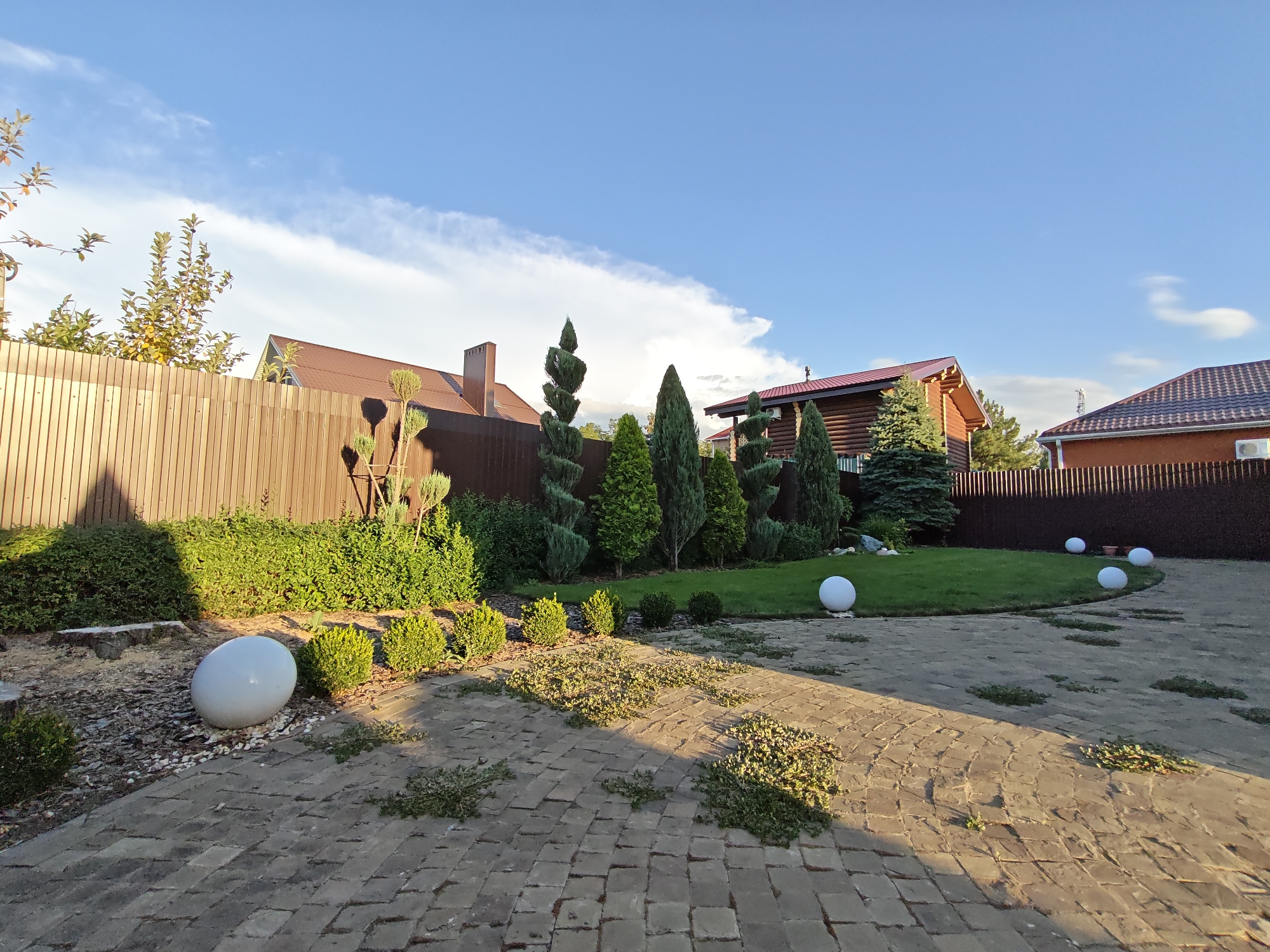 The image depicts a suburban backyard in Vodopadnyy, Russia. The scene is characterized by a well-maintained garden area with a variety of sculpted evergreen trees and neatly trimmed bushes. A paved pathway, made of rectangular bricks, curves through the foreground, with scattered patches of grass growing between the stones.
In the middle ground, a lush green lawn is bordered by a dark brown wooden fence. Several large, spherical white garden lights are strategically placed on the lawn and around the pathway. In the background, two distinct houses are visible. One house appears to be constructed of wood, with a prominent chimney and a red-tiled roof. Adjacent to it is another house with a red-tiled roof and brick exterior. The sky above is a vibrant blue with scattered white clouds, suggesting it is daytime, likely late afternoon or early evening due to the long shadows cast across the scene.
There are no people or animals visible in the image. The overall impression is one of a peaceful and orderly residential setting.