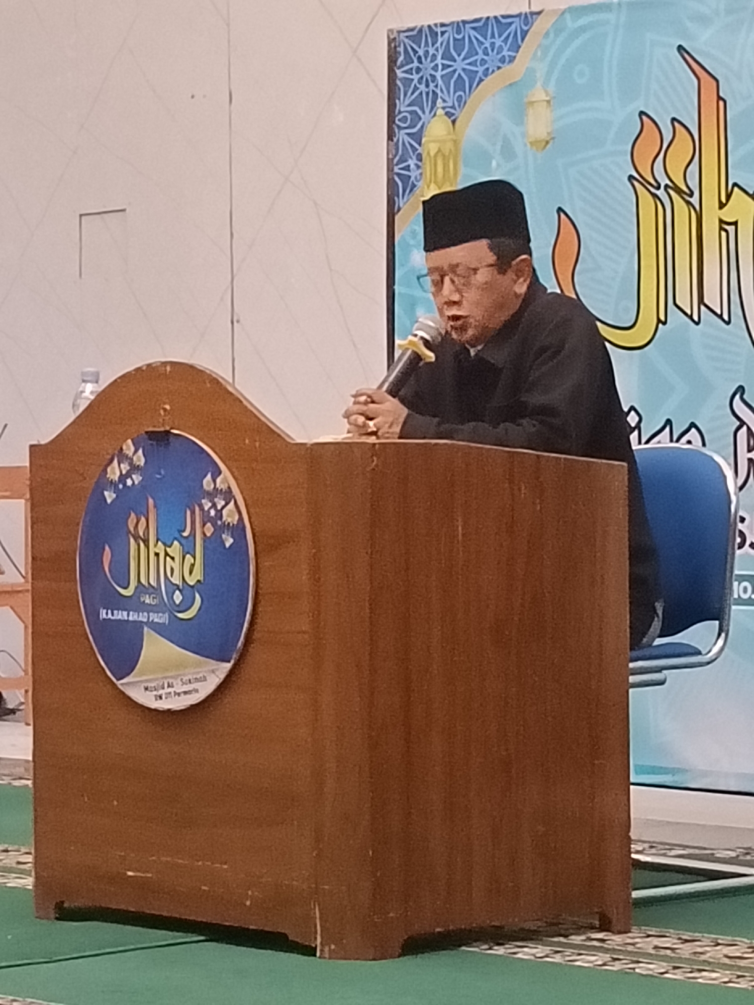A man wearing a black cap and glasses is speaking into a microphone while standing behind a wooden podium. The podium has a circular emblem on its front, featuring Arabic calligraphy and floral motifs, with the words "PAGI" and "(KAJIAN SHAD PAGI)" visible. Behind the man is a backdrop with a blue pattern and a large, stylized yellow and orange Arabic inscription, along with decorative lanterns. A blue chair is visible to the right of the podium. The scene appears to be an indoor event, possibly a religious or educational gathering. The man is wearing a black long-sleeved shirt. His hands are clasped on top of the podium. The backdrop suggests the event is taking place in Kediri City, Indonesia, indicated by the text "Masjid Al-Sakinah DW DTT Parewew".